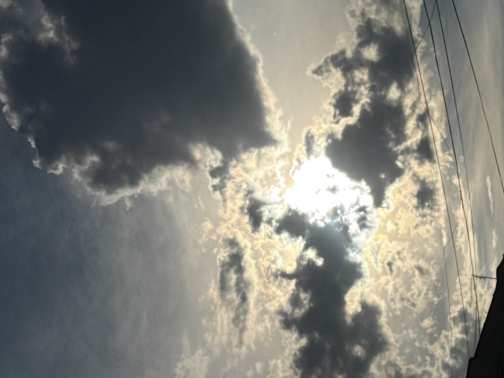 The image captures a cloudy sky with the sun partially obscured by dark clouds. The sun's rays create a bright glow, illuminating some of the lighter clouds. In the foreground, there are power lines stretching across the frame, and the edge of a dark building is visible at the bottom left. The scene appears to be an outdoor setting, likely in the city of Abovyan, Armenia, given the presence of urban infrastructure like power lines and buildings, against a backdrop of a dramatic sky. No people are visible in the frame, and no specific actions or interactions are depicted.