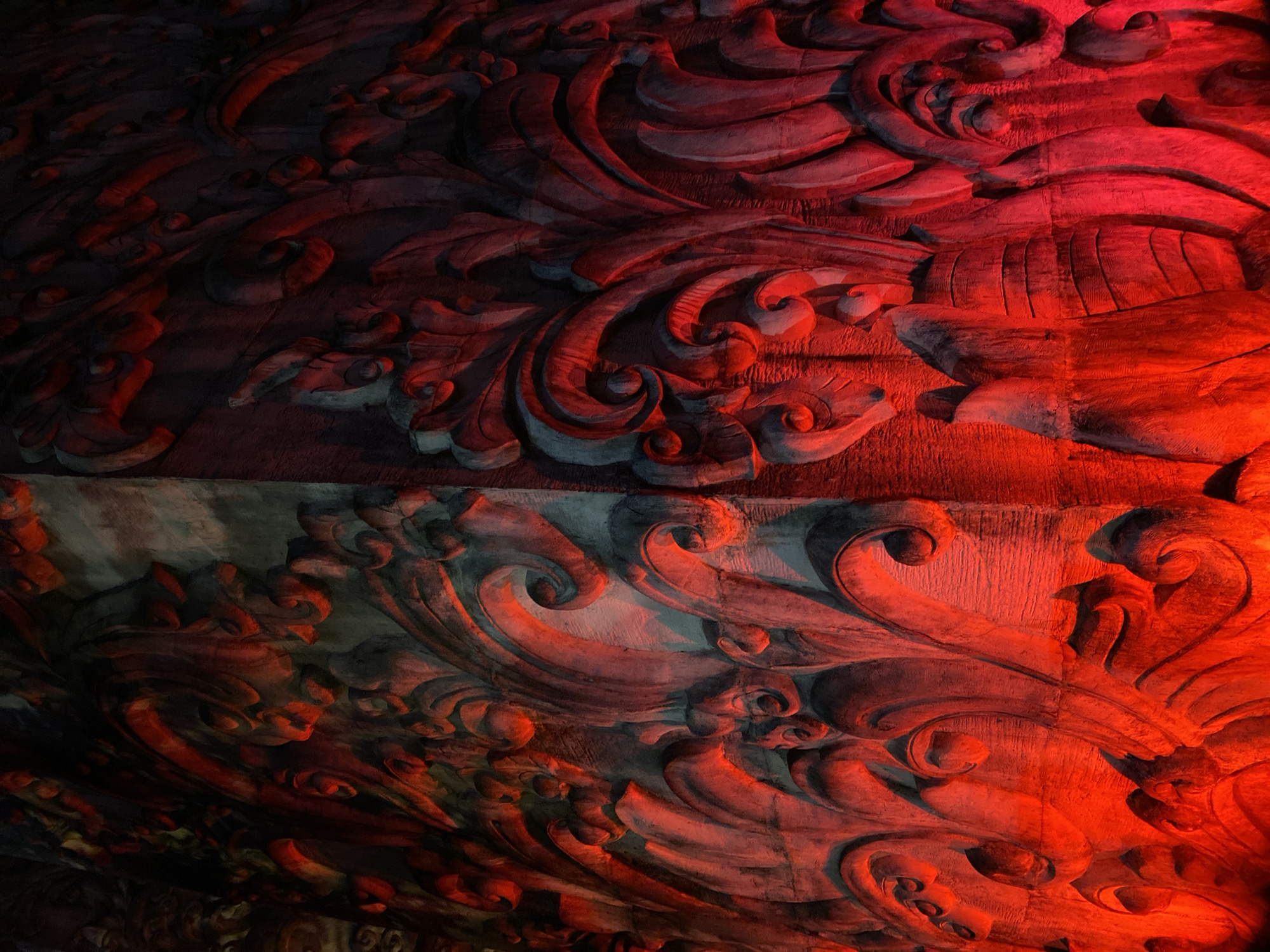 This is a close-up, abstract shot of ornate carvings, likely on a wooden surface, illuminated with a dramatic red light. The carvings feature flowing, organic patterns reminiscent of acanthus leaves and swirling floral motifs. The strong red lighting creates deep shadows and highlights the texture of the material, giving it a dramatic and somewhat mysterious appearance. The lighting is uneven, with brighter red tones on the right side of the image and darker, cooler tones on the left, suggesting a focused light source. The texture of the carvings appears rough and weathered, indicating age or a deliberately rustic finish. The scale is difficult to determine precisely, but the detail suggests it is a significant piece of decorative work. There are no people, animals, or identifiable objects other than the carvings themselves. The scene is indoors or under a covered area, given the artificial lighting. The emotions evoked are dramatic and perhaps spiritual, due to the intense red lighting and the intricate, symbolic nature of the carvings. There is no visible text in the image. The context provided places this in Kuta Selatan, Indonesia, which might suggest traditional Balinese artistry or architectural detail.