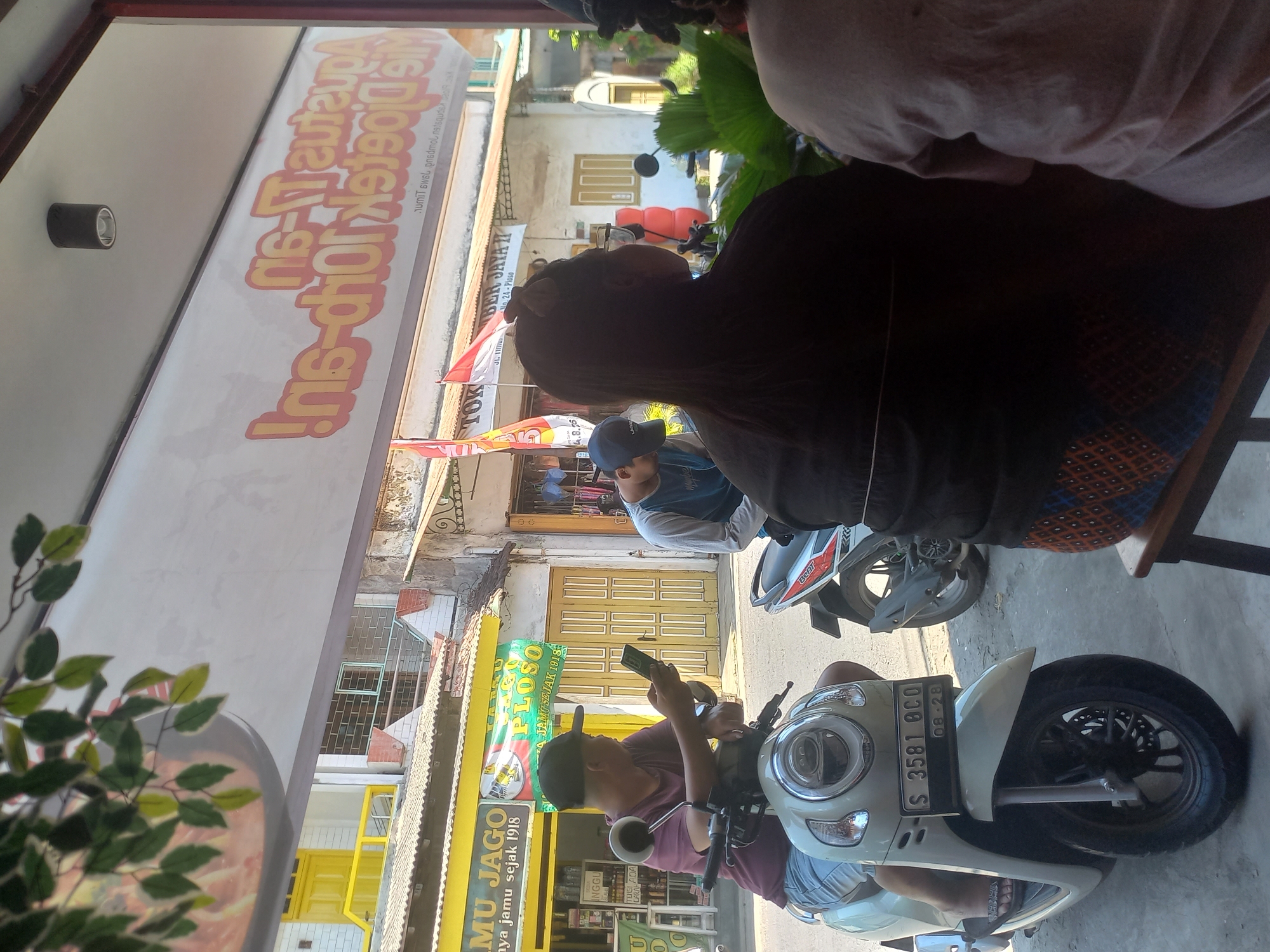 The image depicts a street scene in Ploso, Indonesia, likely in front of a small business. In the foreground, a person is seated on a light-colored scooter, holding a mobile phone. The scooter's license plate reads "S 3581 OCO 08-28." To the right of this person, another individual is seated, facing away from the camera. Behind them, a man wearing a cap is seated on another scooter, also looking down at his phone.
The background reveals storefronts with signage. One sign, visible in the upper portion of the image, is a large banner with text in orange and red, appearing to advertise a food establishment. Below this, another sign reads "JAMU JAGO" and "JAMU SEJAK 1918." Further down the street, more buildings and businesses are visible, with yellow doors and corrugated metal roofs. Red and white flags are also present, possibly indicating a local celebration or business. The environment suggests a warm, sunny day.