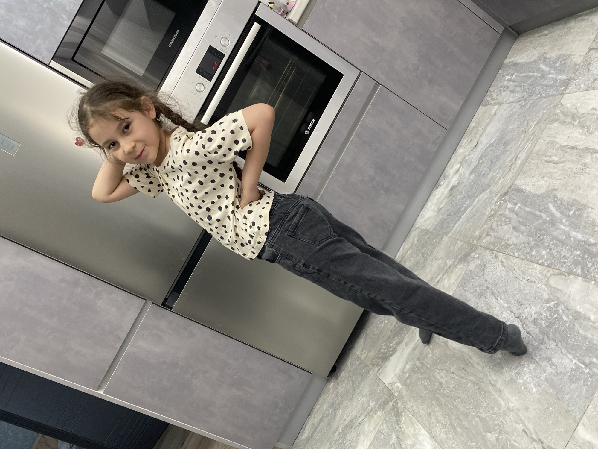 A young girl is pictured standing and posing in a modern kitchen, likely in Izhevsk, Russia. She has dark hair styled in two braids and is wearing a light-colored short-sleeved t-shirt with black polka dots and dark-wash jeans. Her feet are covered in dark socks or are bare. She stands with her hands on her hips, one arm bent with her hand behind her head, and is looking directly at the camera with a subtle, playful smile.
The kitchen features contemporary design elements, including light grey, textured, flat-panel cabinets and stainless steel or highly reflective cabinet surfaces. Built into the cabinetry are a Grundig microwave oven and a Bosch electric oven. The digital display on the Bosch oven clearly shows "15:00," indicating the time of day. The floor is covered with light grey, marble or stone-look tiles. The photograph is taken from a slightly high angle and is tilted, giving the impression that the girl and the kitchen are leaning.
