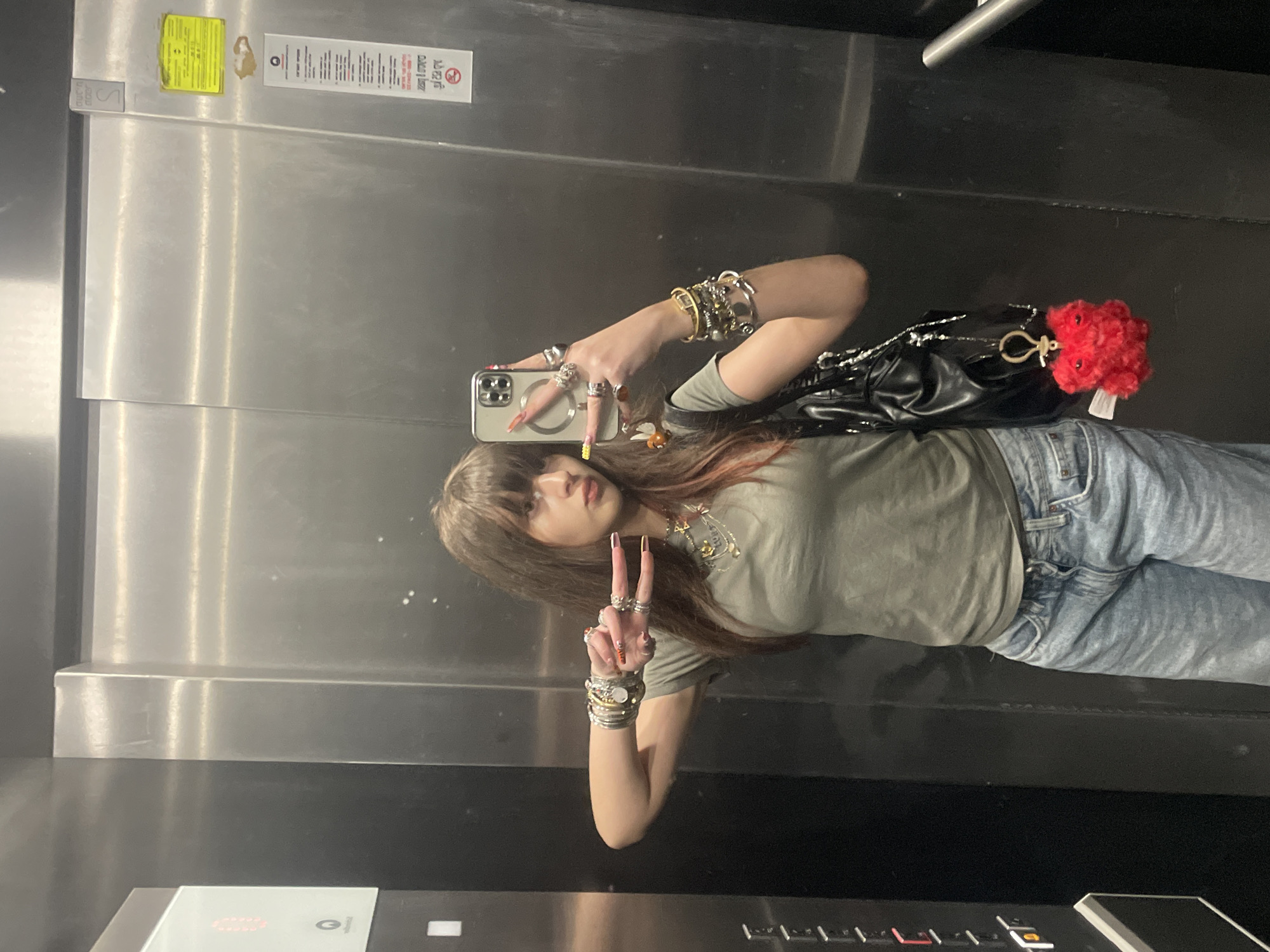 A woman takes a selfie in an elevator. She wears light-wash jeans, a green t-shirt, and many bracelets and rings. She is making a peace sign with her fingers. She has a black purse with a red pom-pom hanging on her shoulder. The elevator has stainless steel walls with various signs and labels. The location is Raanana, Israel.
