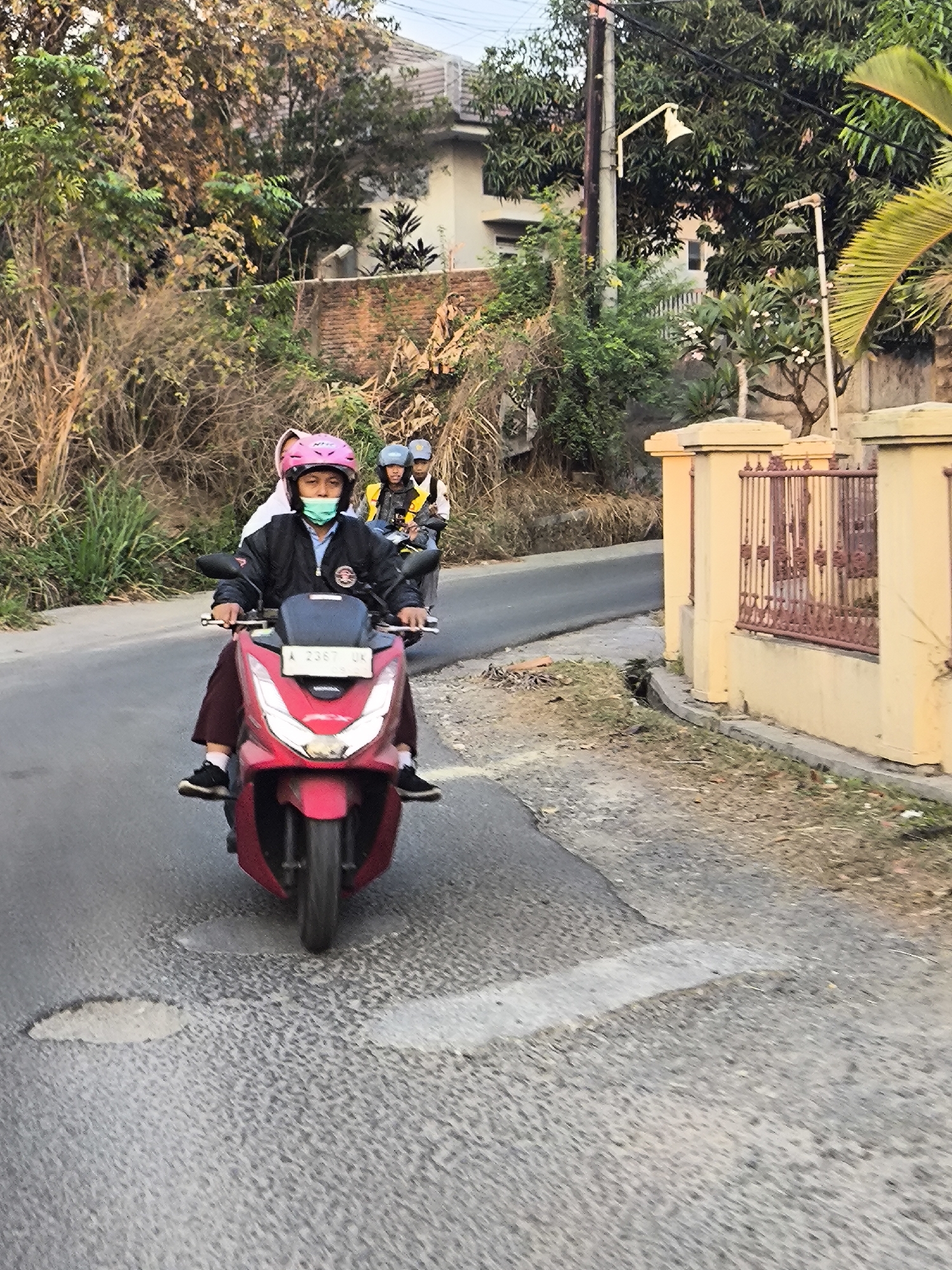 The image depicts a group of individuals on motorcycles traveling along a paved road in Cilegon, Indonesia. In the foreground, a person wearing a pink helmet and a light blue face mask rides a red scooter. This individual is wearing a dark jacket and dark pants. Behind them, another scooter carries two passengers, both wearing helmets and face masks. The road appears to be in a somewhat rural or suburban area, with vegetation and trees lining the sides, and a yellow-colored fence with a decorative metal railing visible on the right. The sky suggests it is daytime, possibly late afternoon due to the lighting. The scene is captured from a slightly elevated perspective, looking down at the road.