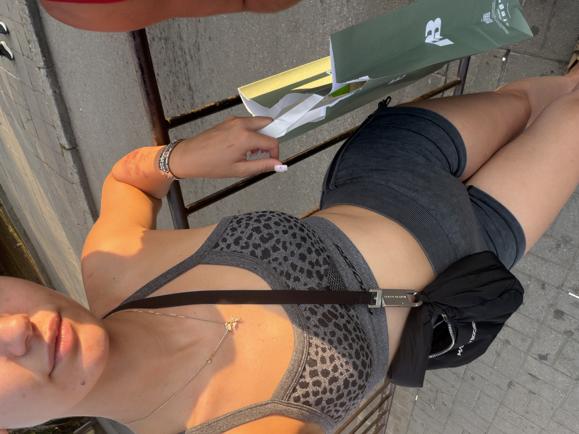 An unidentifiable person is positioned outdoors, likely in a city, with warm, golden light suggesting sunset or sunrise. They are leaning on a railing, with their right arm resting on it. The person is wearing a grey leopard-print sports bra, dark grey athletic shorts, and a black crossbody bag. A silver necklace with a small pendant is visible. A green shopping bag is held in their right hand. In the background, a paved walkway and a street with some distant figures are visible. The overall impression is of a casual, outdoor setting.