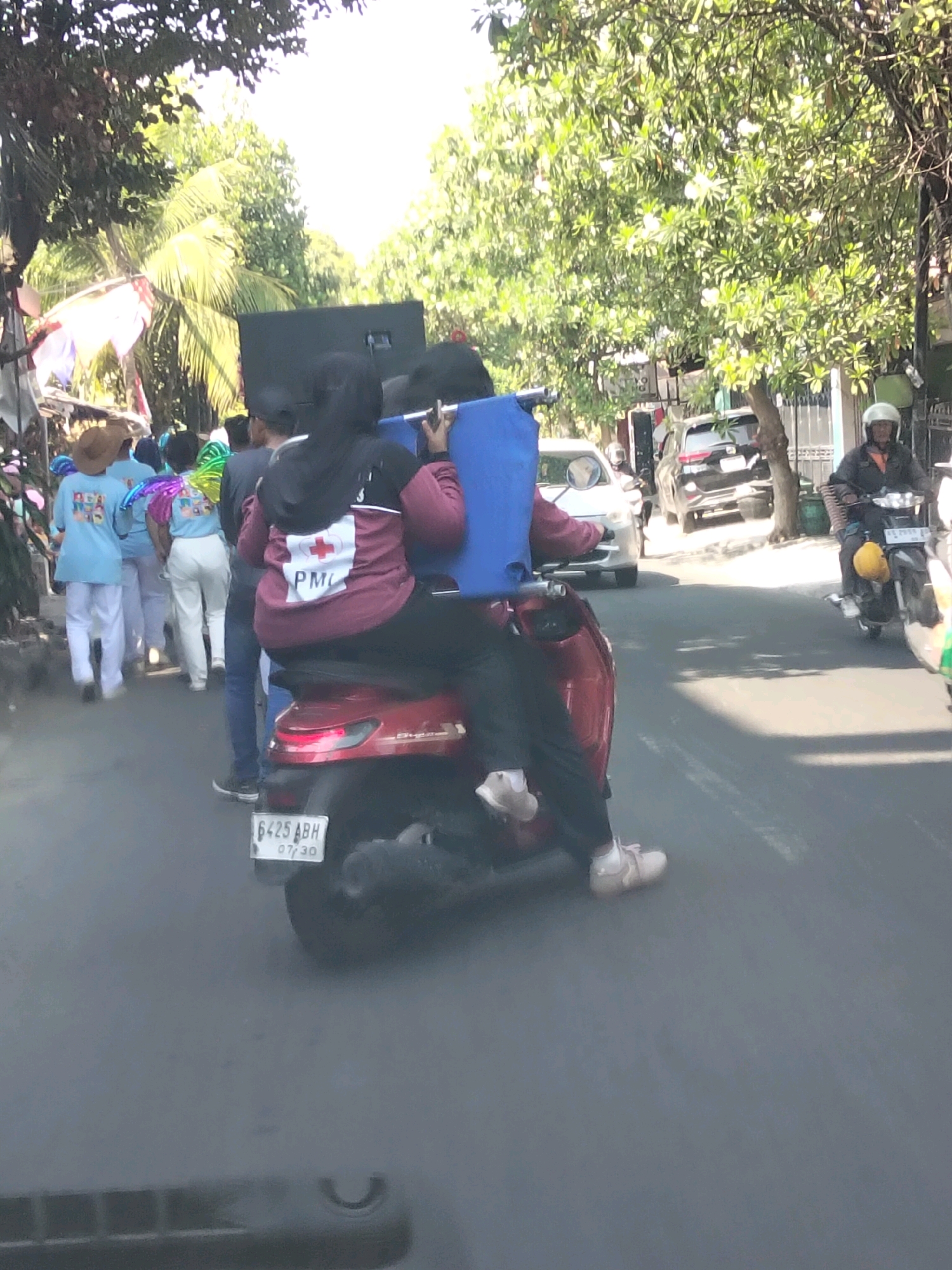 The image depicts a street scene in Kediri City, Indonesia, on a sunny day. In the foreground, a red scooter is being ridden by a person wearing dark pants and light-colored shoes. A blue carrier is attached to the back of the scooter. Behind the scooter rider, another person wearing a maroon jacket with a white cross and the letters "PMC" is visible. The scooter has a license plate that reads "6425 ABH 07/30".
Several other people are walking on the sidewalk to the left of the scooter. One person wears a straw hat and a light blue t-shirt, while another has colorful wings attached to their back and a similar blue shirt. They appear to be part of a procession or event.
In the background, several cars are present on the street, including a silver sedan and a dark SUV. A person wearing a white helmet is riding a motorcycle further down the road. Trees line the street, casting shadows and creating a natural canopy over parts of the scene. Buildings and fences are visible along the side of the road. The overall atmosphere suggests a busy street during a daytime event.