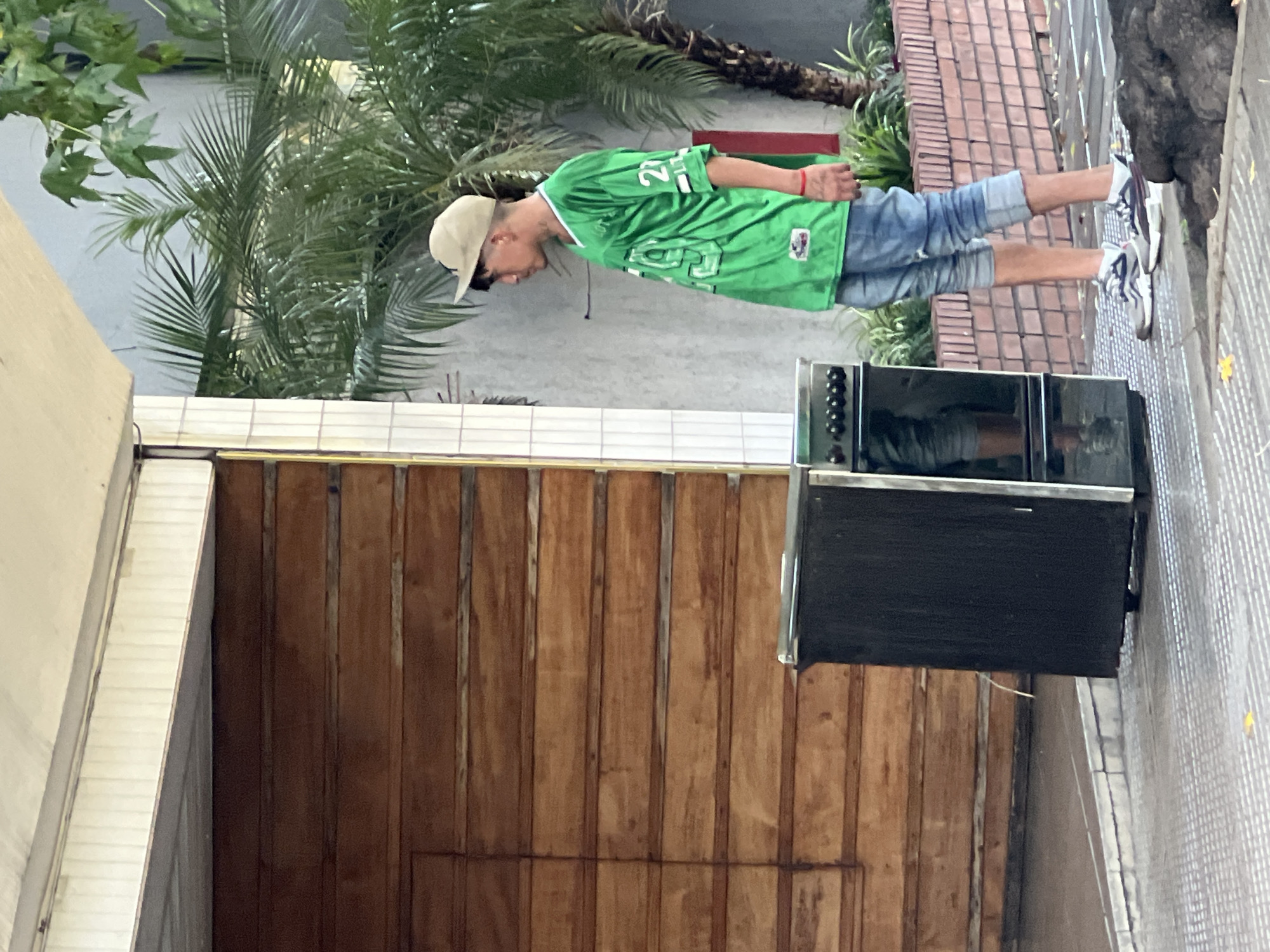 A vertical, medium-shot depicts a man standing outside in what appears to be Buenos Aires, Argentina. The man is wearing a green sports jersey with the number 22, blue jean shorts, white sneakers, and a tan baseball cap. He stands beside an old oven. Behind the man is a wooden door, various plants, and brick pavers.