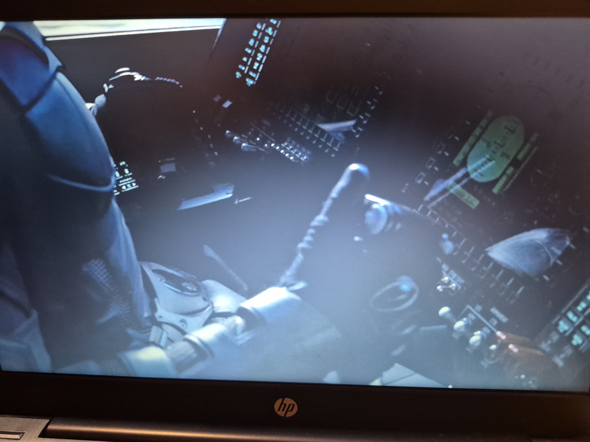 The image is a close-up, slightly blurred photograph of a person in a futuristic cockpit. The person is wearing a dark blue suit, with a white and silver mechanical arm or glove visible on the left side of the frame. The cockpit is filled with glowing screens and controls. On the right side of the frame, a green circular display with some text is visible. The overall color palette is dark with bright, glowing accents from the displays. The image is captured from an angle that suggests the viewer is looking over the shoulder of the person in the suit. The bottom of the image shows the edge of a laptop with an HP logo. The photograph does not contain any recognizable elements that would allow for the identification of its location in Sandwell, United Kingdom.