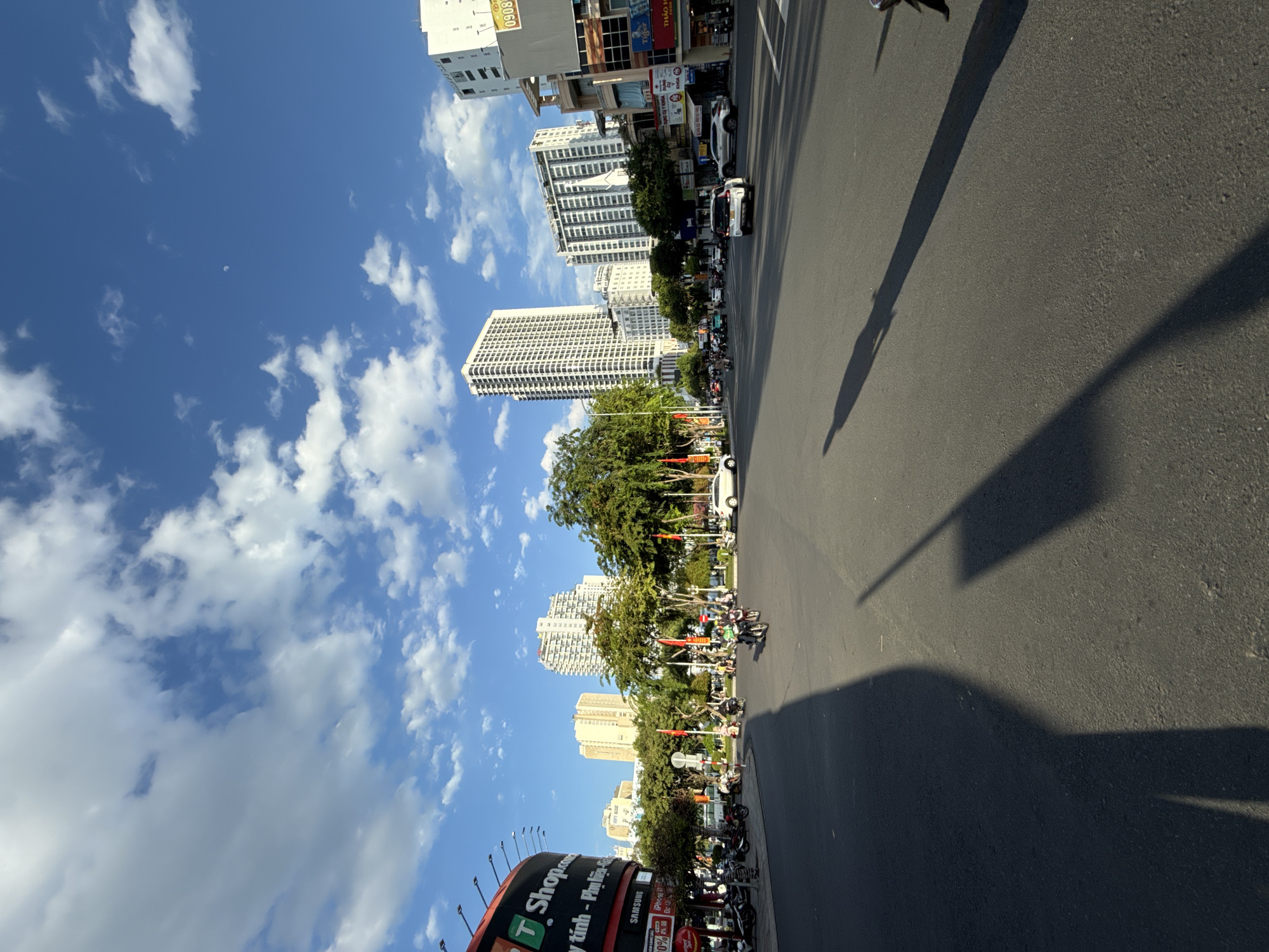 This image captures a wide-angle view of a street intersection in Nha Trang, Vietnam, under a bright blue sky with scattered white clouds. The foreground is dominated by an asphalt road, with distinct shadows of people and possibly a lamppost cast across it, indicating it is daytime.
In the middle ground, the intersection is busy with traffic, primarily motorcycles and a few cars. Several motorcycles are visible on the road, some with riders. A white sedan is seen turning, and another white car is parked or moving slowly on the right side of the frame. Pedestrians are present on the sidewalks and near the intersection. Trees line the sides of the road, providing shade.
The background features a cityscape with several tall buildings, characteristic of urban development in Nha Trang. These include modern high-rise apartment or office buildings and some lower structures. A large, circular building with signage for "T Shop.com" is prominently visible on the left side of the frame. Vietnamese flags are displayed on poles along the street. The overall scene depicts a typical day in a bustling Vietnamese city.