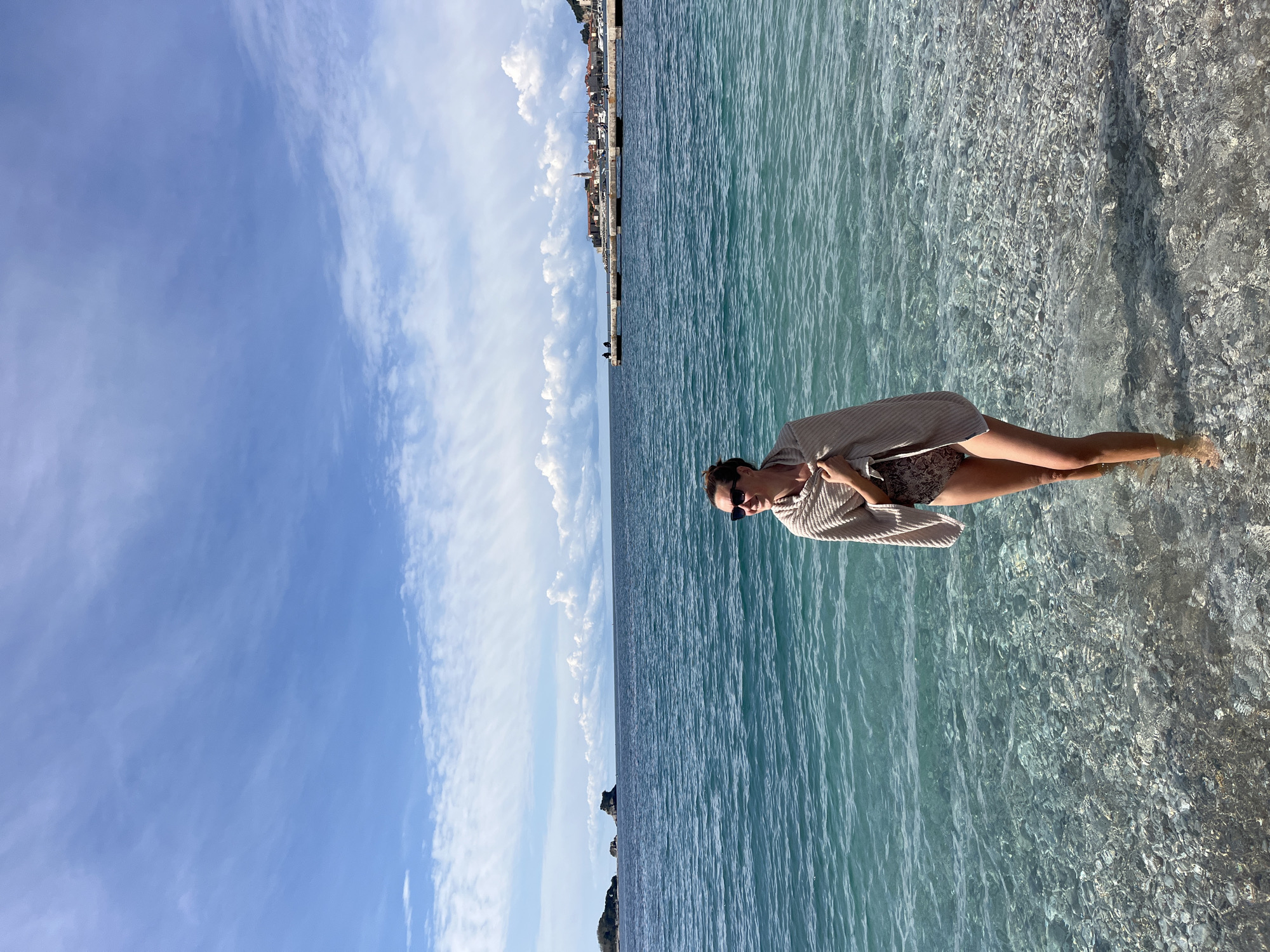 A woman stands in shallow, clear turquoise water at a sunny coastal location, likely in Budva, Montenegro. The water, calm and rippled, reveals pebbles and rocks on the seabed where her legs are submerged up to the calves. She is wearing a patterned swimsuit bottom and a light-colored, possibly knitted, cover-up or top, paired with sunglasses. Her hair is pulled back, and she is looking away from the camera, towards the left of the frame.
The horizon line stretches across the image, separating the sea from a bright blue sky adorned with scattered white clouds. In the distance, along the coastline, a city or town with numerous buildings is visible, suggesting a populated area. Further out, small islands or rock formations emerge from the water. The time of day appears to be bright daylight, with clear sunny weather. There is no visible text in the image.