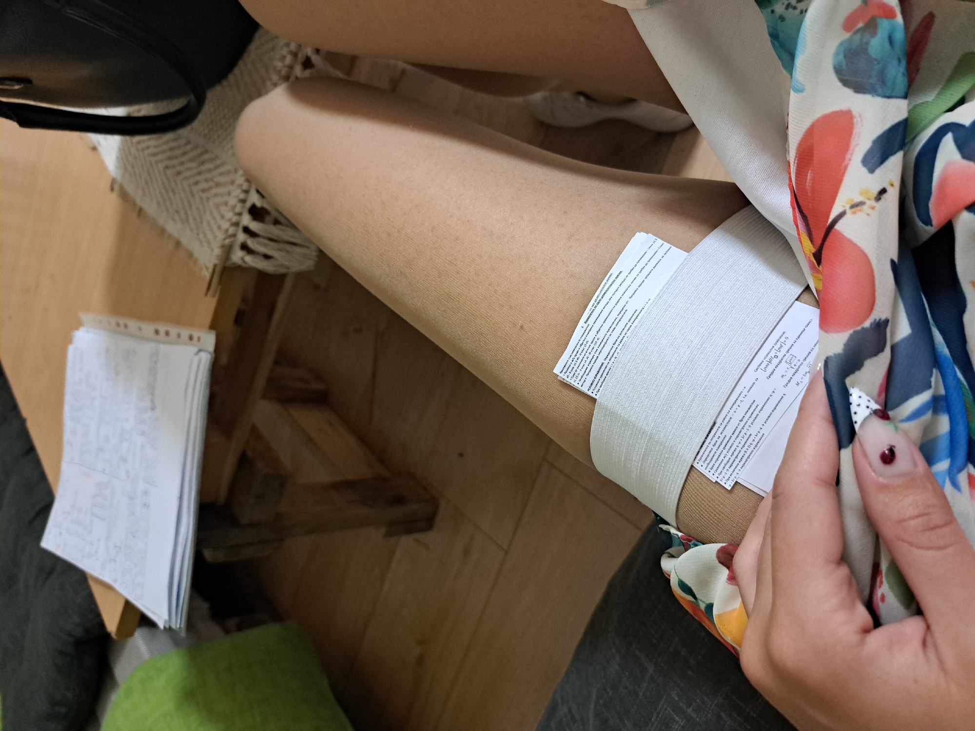 This indoor shot shows a person sitting with papers wrapped around their leg. The leg is covered in what appears to be a tan-colored stocking. A white bandage is wrapped around the upper part of the leg, with two papers stuck to it. The paper has Cyrillic text on it. The hand of the person holds a floral scarf. There are papers on the floor to the left of the leg, as well as a black purse. The floor and a wooden chair are visible. There is no indication of this being located in Sofia, Bulgaria.