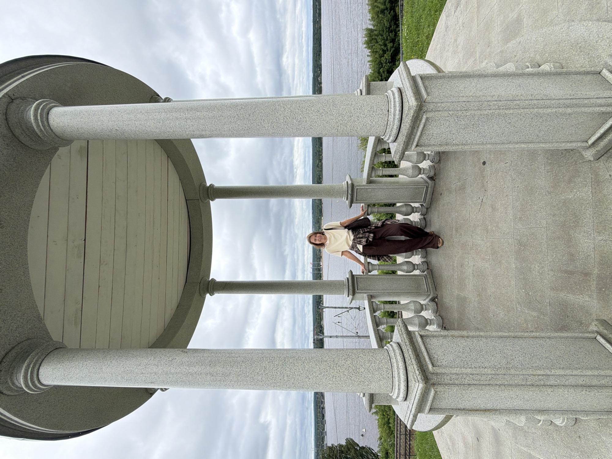 A woman stands inside a white-columned gazebo, leaning against the railing. She is wearing a light-colored t-shirt, brown pants, and a plaid shirt tied around her waist. She carries a dark bag over her shoulder. The gazebo overlooks a wide river with a cityscape on the opposite bank. The sky is overcast. The gazebo is located at the Razgulyay embankment in Perm, Russia.