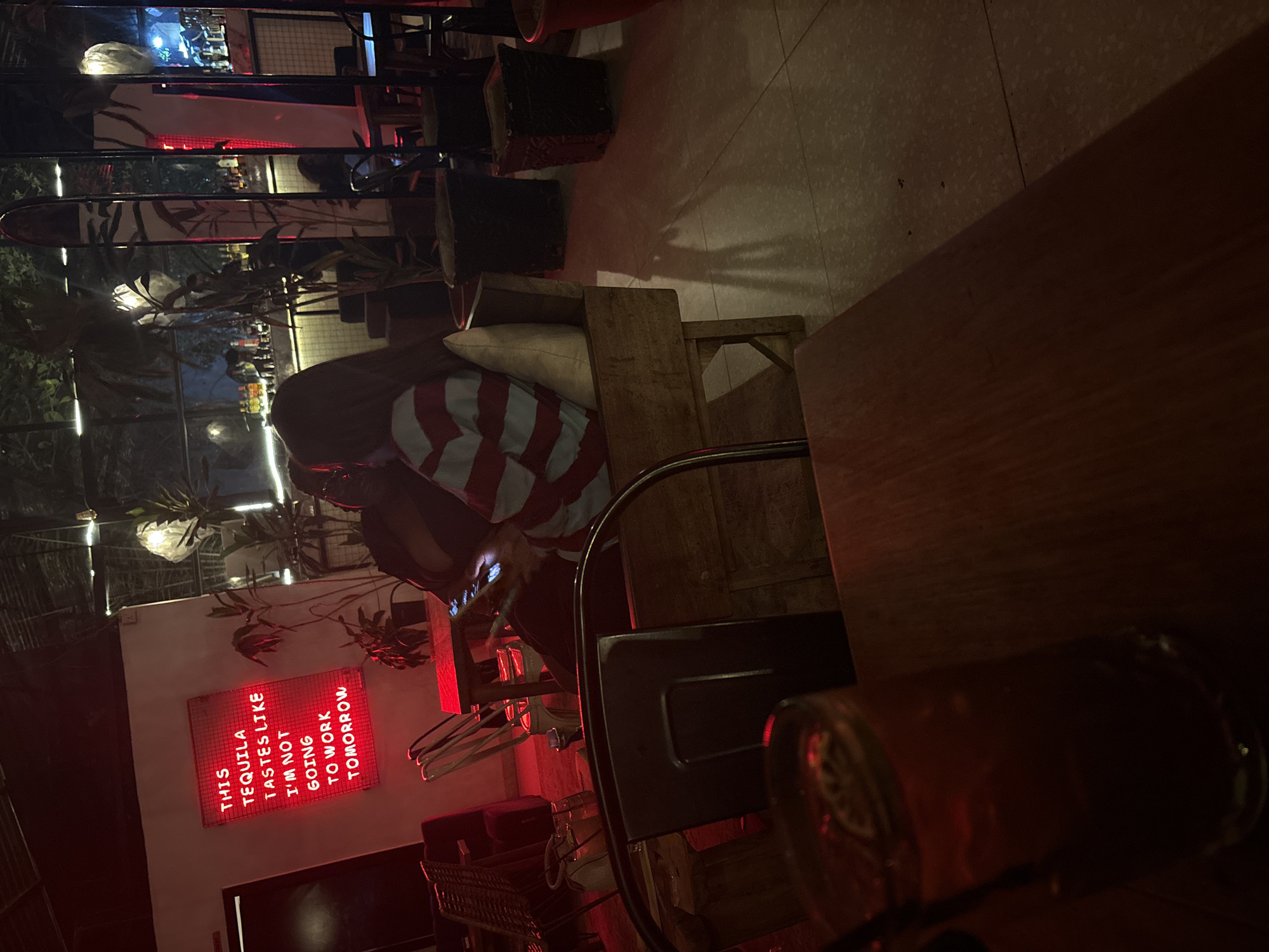 A person sits at a wooden table, illuminated by dim, reddish light. A glass of amber liquid sits in the foreground. The person is looking at their phone. Behind them, a neon sign reads, "THIS TEQUILA TASTES LIKE I'M NOT GOING TO WORK TOMORROW." Based on the decor and style, this is likely a bar or restaurant. The specific location in Abuja, Nigeria is not discernible from the image.