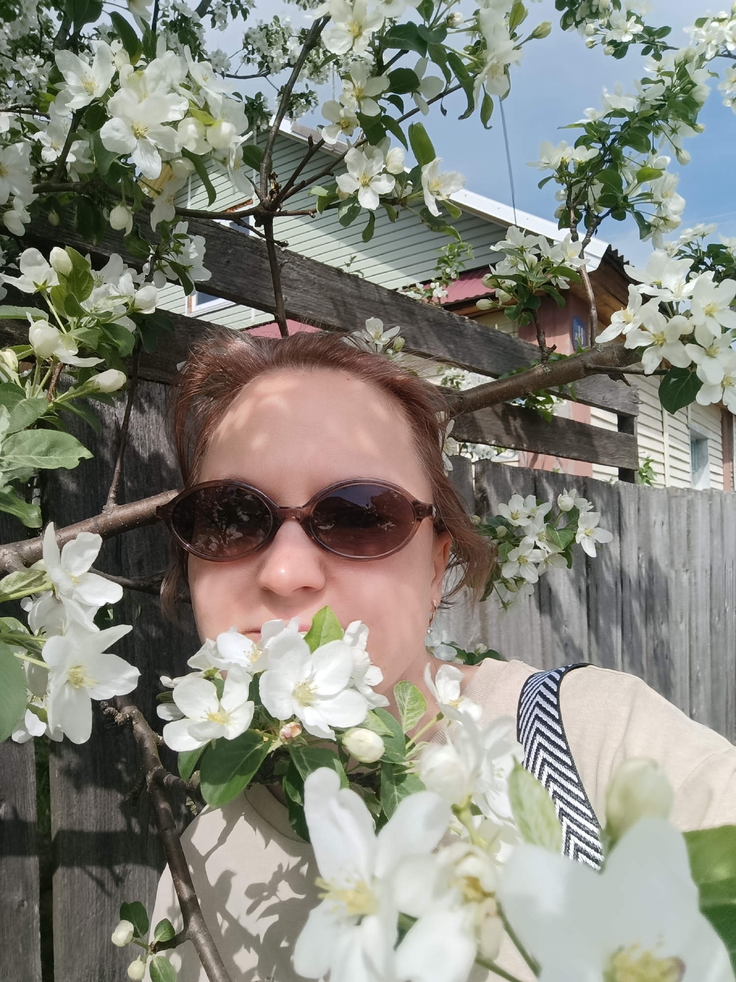 A woman is posing for a selfie in front of a wooden fence. The frame is filled with branches of a flowering tree with white blossoms. She is wearing brown sunglasses and a light-colored shirt with a dark-and-white striped strap. Behind the fence, portions of houses are visible, including a light green house with white trim and a house with a red roof. The image appears to be taken outdoors on a sunny day in Dubovaya, Russia.