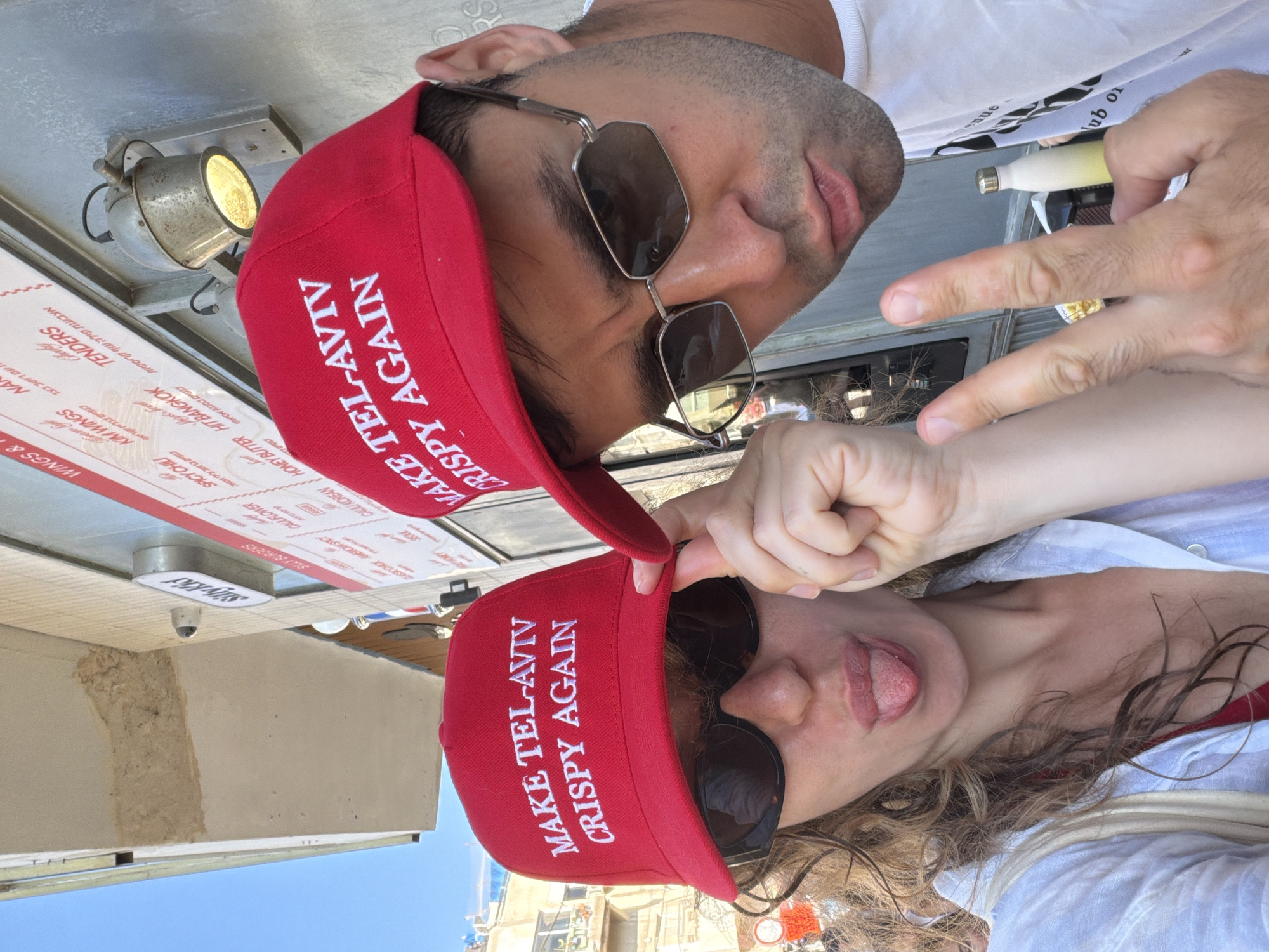 The image shows two people wearing red hats with the text "MAKE TEL-AVIV CRISPY AGAIN" in white. The person on the left is a woman with curly brown hair and sunglasses, sticking her tongue out and making a peace sign with her hand. She is wearing a white shirt over a red bikini top. The person on the right is a man with dark hair and sunglasses, also making a peace sign. He is wearing a white t-shirt. In the background, there is a building with a menu board on the wall and an outdoor light fixture. The menu board lists various food items, including "WINGS," "TENDERS," "SPICY CHILI," and "HONEY BUTTER." A sign above the menu reads "SIVKID." The scene appears to be an outdoor food establishment in Tel Aviv, Israel, as indicated by the text on the hats. The overall atmosphere is casual and playful.