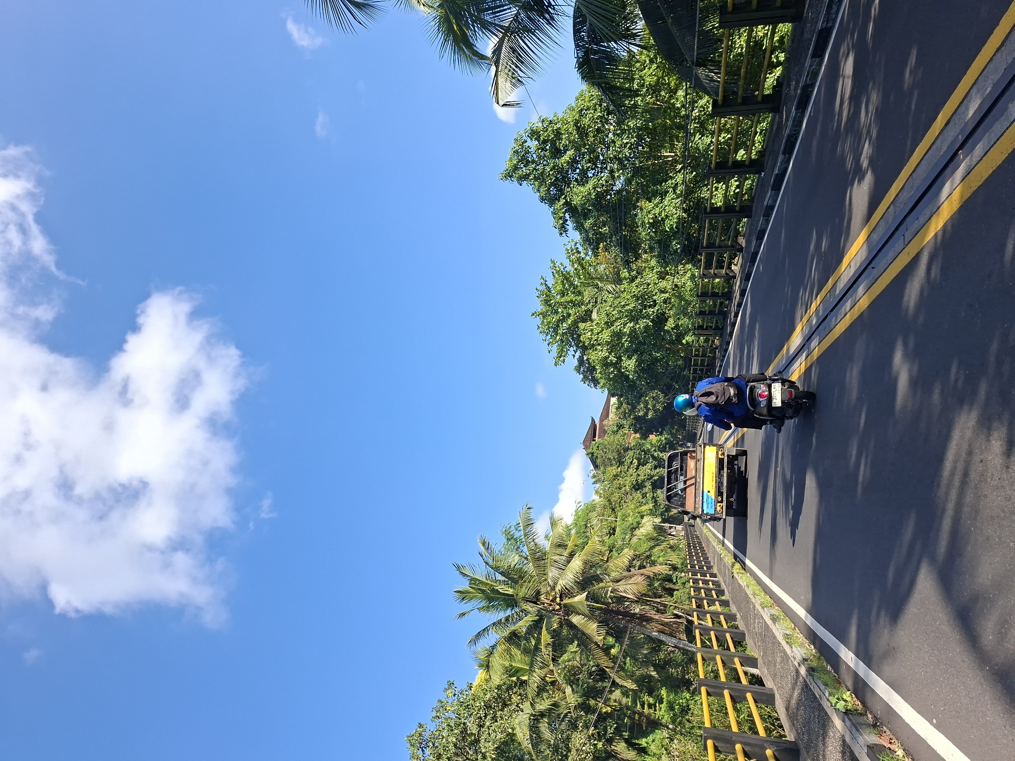 An eye-level, medium shot captures a sunny day on a road that appears to be in or near Banjarangkan, Indonesia, given the presence of lush tropical foliage. The road is paved with asphalt and marked with two yellow lines down the center, with a white line on the right edge. On the left side of the road, a walkway with a yellow metal railing runs alongside, bordered by vegetation and trees, including a prominent palm tree. On the right side, the road is also bordered by trees and dense foliage.
In the foreground, a person wearing a blue shirt, a blue helmet, and a backpack rides a scooter, heading away from the camera. Behind the scooter, a small yellow truck with a blue and white load bed is also traveling in the same direction. The sky is a clear, bright blue with a few scattered white clouds. In the background, several trees are visible, along with a hint of a traditional Balinese-style building with a dark, sloping roof. Shadows from the trees are cast across the road.