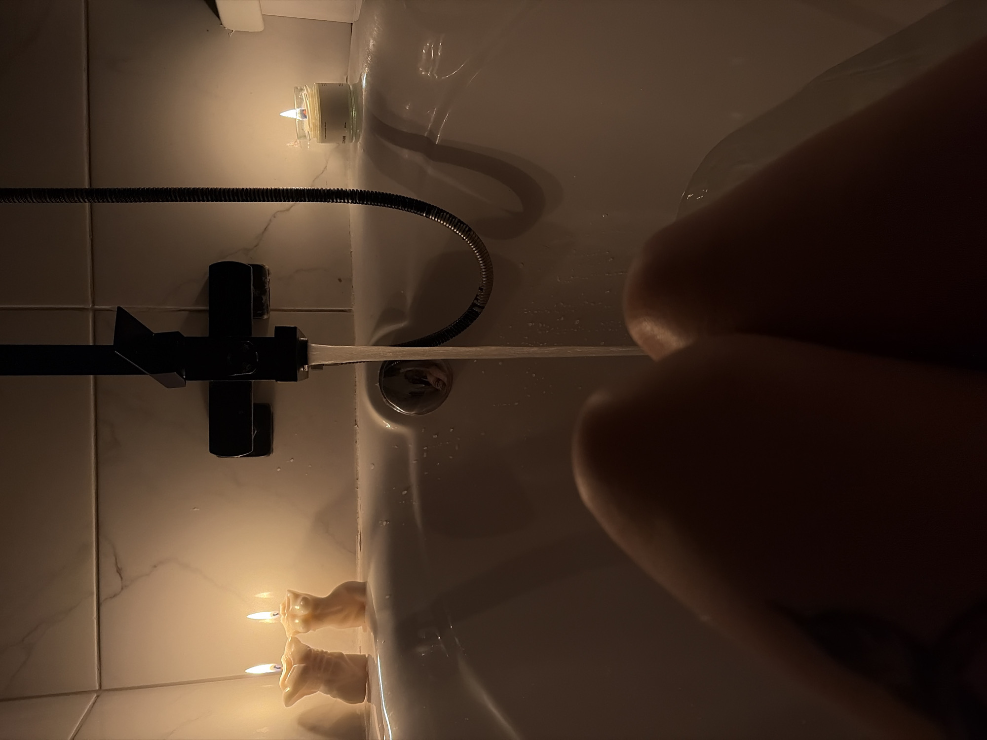 A dimly lit bathroom scene features a white bathtub with running water from a black faucet. Two candles are lit on one end of the tub, while another candle is positioned near the showerhead. A person's body is partially visible in the tub. The bathroom walls are tiled with a marble-like pattern. The scene appears to be indoors. The location, Chaikovsky, Russia, cannot be verified from the image.