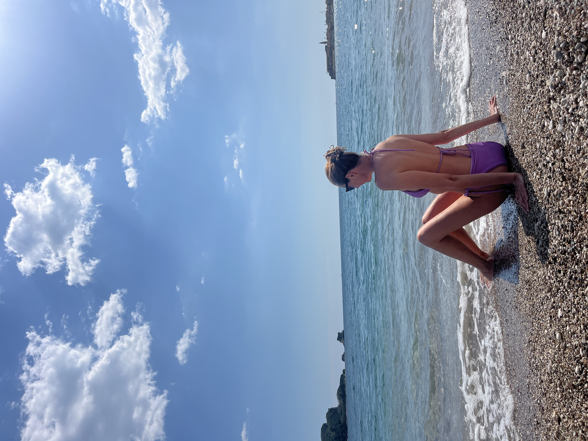 A woman in a purple bikini is kneeling on a pebble beach in Budva, Montenegro. The sea is to her left, and the sky is above. The sky is blue with white clouds. The waves are lapping at the shore. In the distance, there is a building on the horizon.