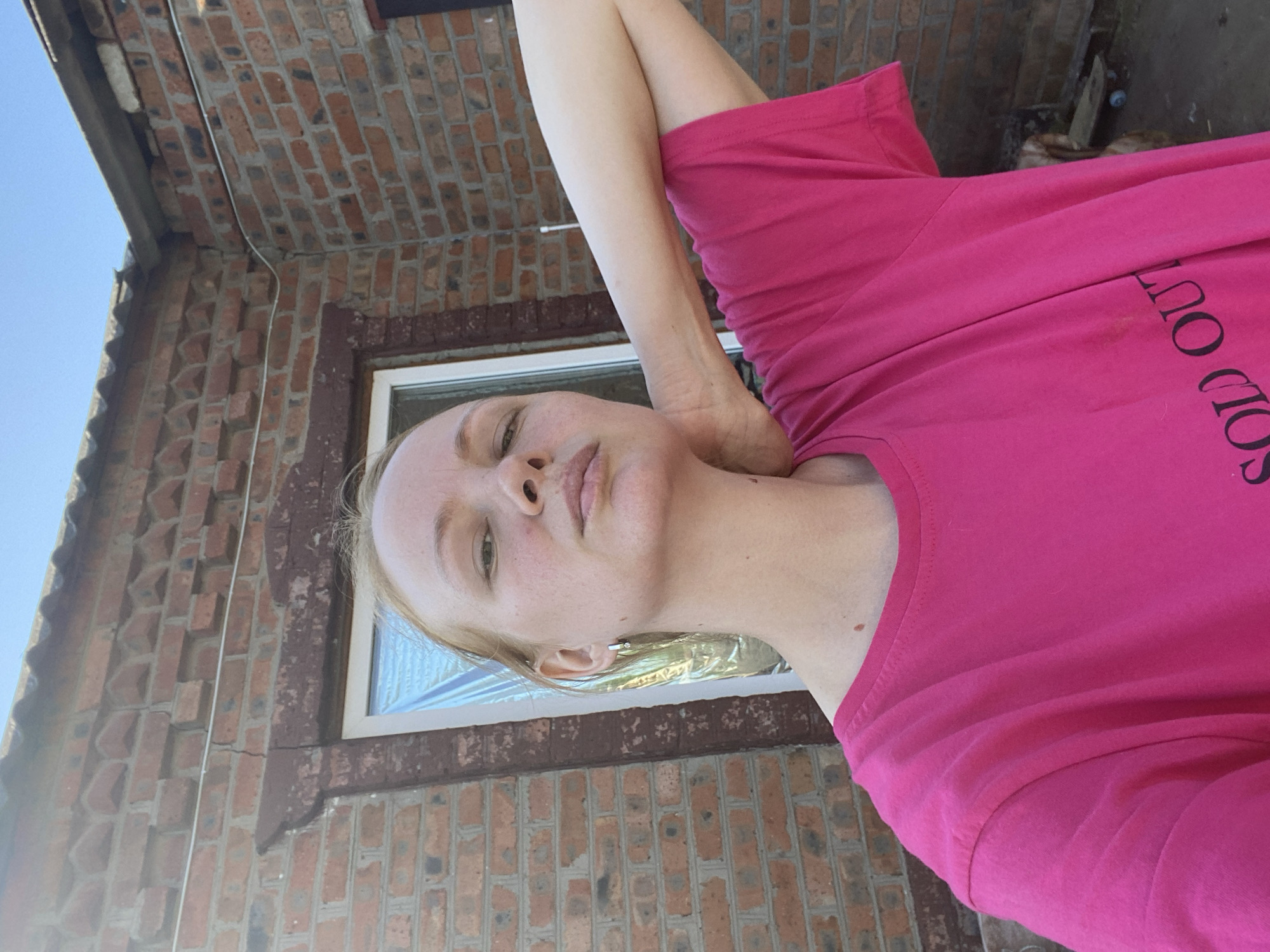 A fair-skinned, fair-haired woman with freckles and light makeup poses for a selfie outdoors in front of a red brick building. She is wearing a bright pink t-shirt with black lettering that reads "SOLD OUT". Her right arm is bent behind her head. The background consists of a brick wall and a window with silver sheeting covering it. A portion of a corrugated roof is visible at the top of the frame. The sky above is clear and blue. The scene is likely in Kisliakovskaya, Russia, given the architectural style of the building.