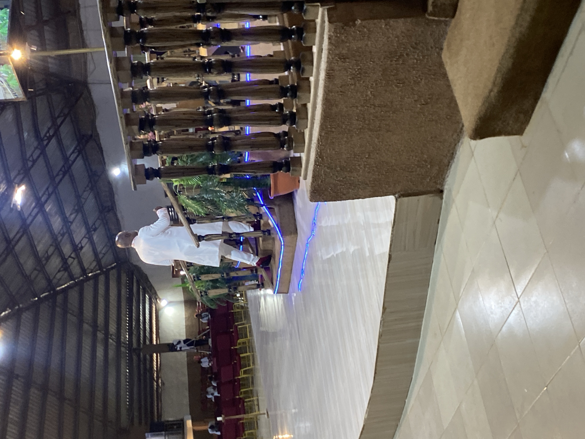 A man in a white robe is ascending a staircase in a large room. The room appears to be a church or auditorium, with rows of red chairs visible in the background. The staircase has a decorative wooden railing and is lit with blue LED lights. The building appears to have a metal roof. This appears to be in Jos, Nigeria.