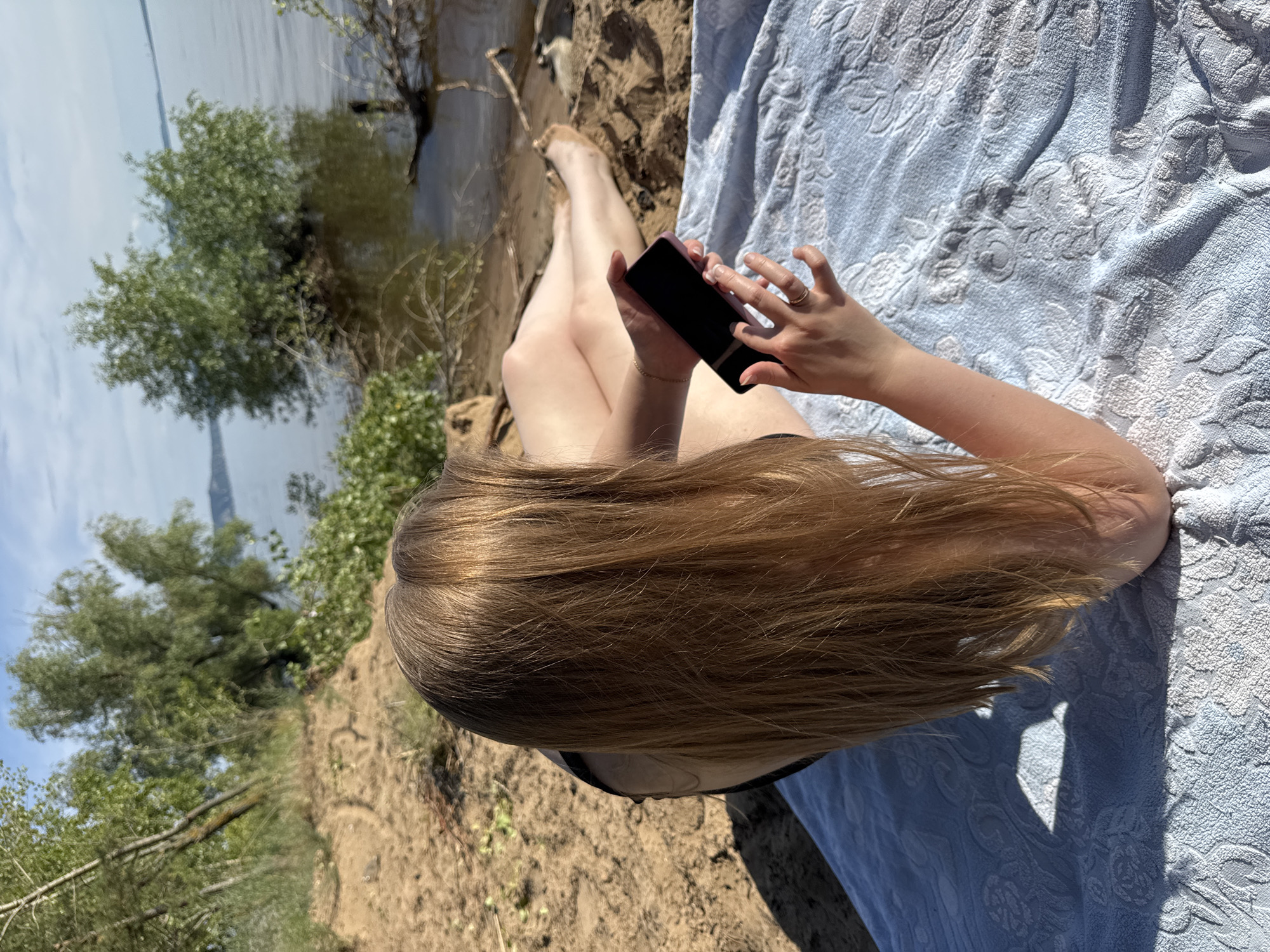 A high-angle, full-body shot captures a person lying on their back on a blue, patterned towel by the water. The person's legs are extended, with their feet toward the water's edge. They are holding a dark-colored smartphone in their hands, with their fingers on the screen, suggesting they are interacting with it. Their long, blonde hair is spread out around their head, falling onto the towel.
The scene is a sandy embankment next to a body of water, likely a lake or river, given the calm surface and the trees lining the opposite bank. The environment appears natural, with trees and dry brush visible. The lighting suggests it's daytime, with bright sunlight casting shadows. The weather appears to be pleasant, suitable for relaxing outdoors. The overall mood is tranquil and relaxed, with the person engrossed in their device while enjoying the natural setting. No text is visible in the image.