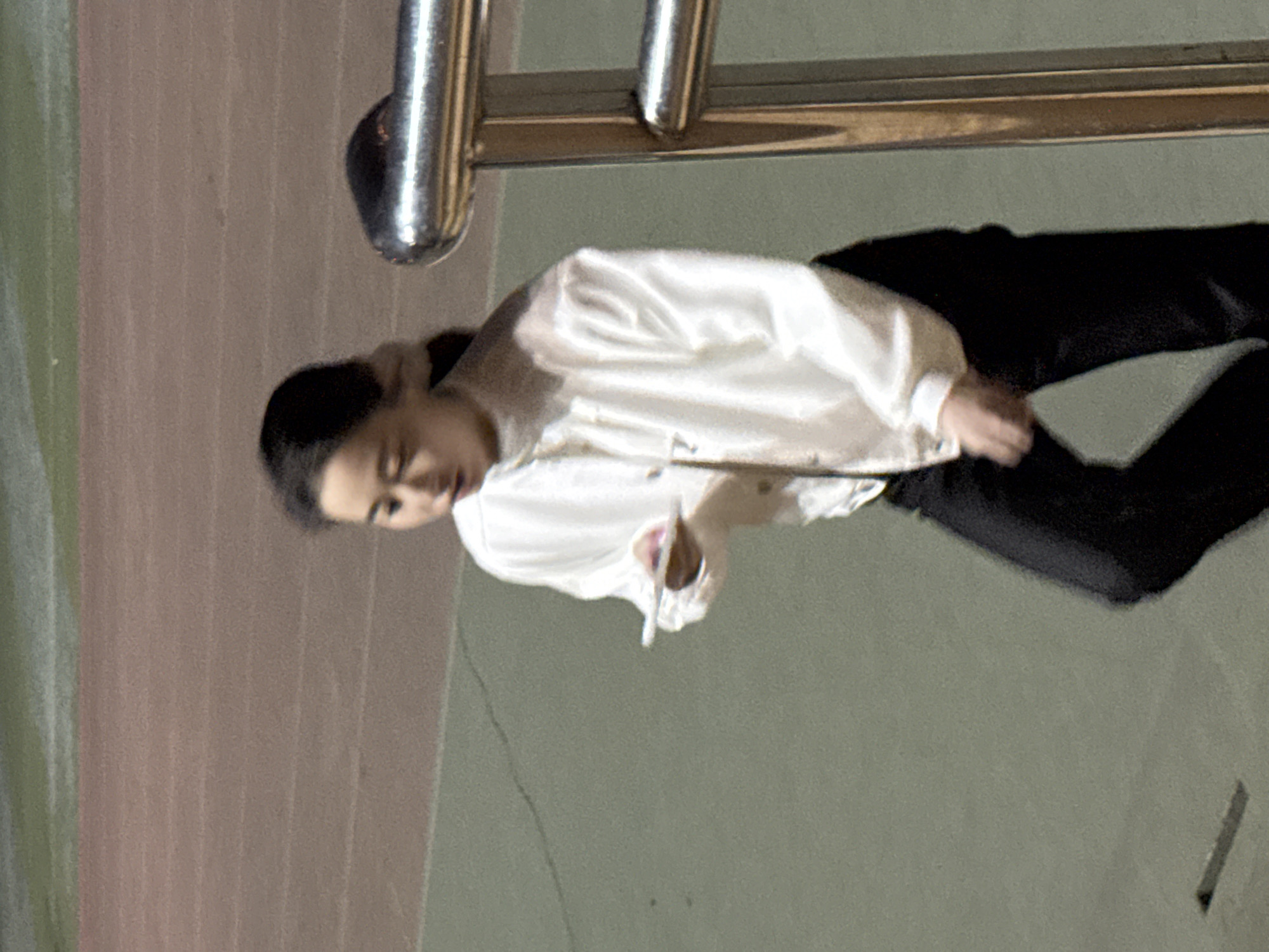 This image, taken in Zhongtun, China, portrays a single person, likely a young woman, standing in what appears to be an indoor or covered outdoor area, possibly a walkway or corridor. The image is currently rotated 90 degrees clockwise.
The subject is wearing a light-colored, long-sleeved top or jacket, possibly white or off-white, and dark-colored trousers. Her dark hair is pulled back. She is looking downwards and slightly to her left, with a focused or neutral expression. Her right hand (viewer's left when viewed correctly) is slightly raised, holding a small, light-colored object, which appears to be a folded piece of paper or a similar item.
In the background, there is a textured wall with subtle vertical stripes in muted brown and possibly green tones. Metallic handrails or barriers are visible in the upper portion of the frame (when correctly oriented), suggesting architectural features. The ground or floor beneath her is a solid, muted green-gray color. The lighting in the scene is dim and diffused, hinting at an evening, nighttime, or artificially lit indoor setting. There is no visible text present in the image.