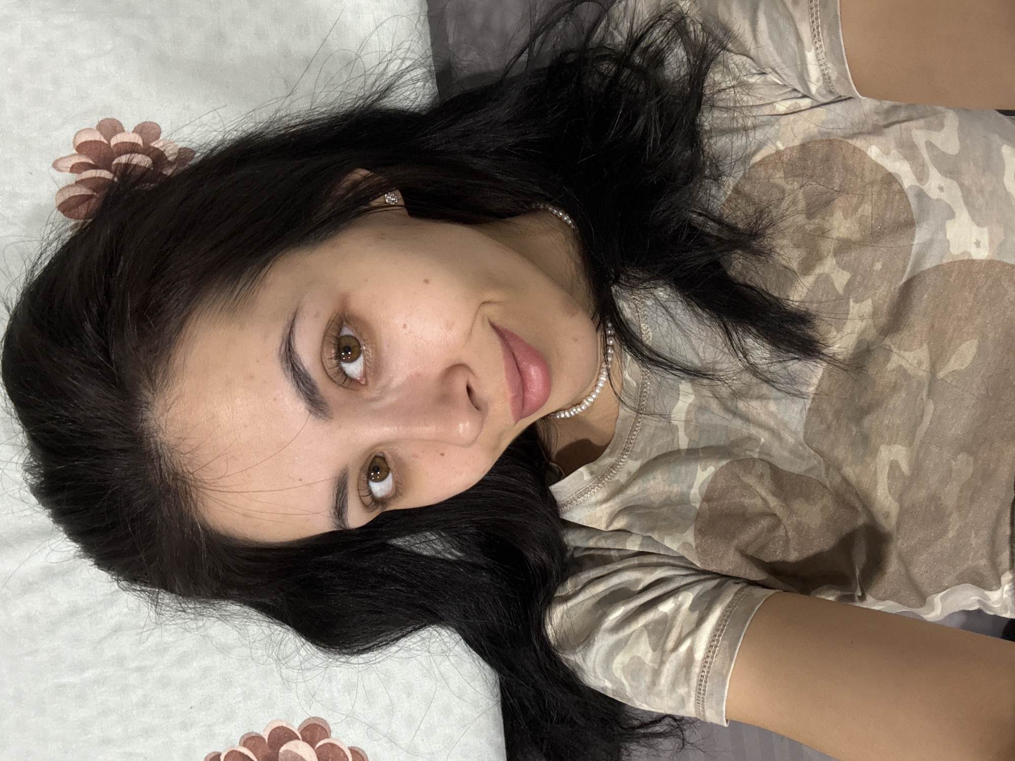 A woman with dark hair and brown eyes is lying down, looking up and to her left. She is wearing a light brown camouflage pattern t-shirt and a pearl necklace. Her head is resting on a patterned pillow with brown floral designs in the corner. The background is out of focus. There is no information available to determine the location in Votkinsk, Russia.
