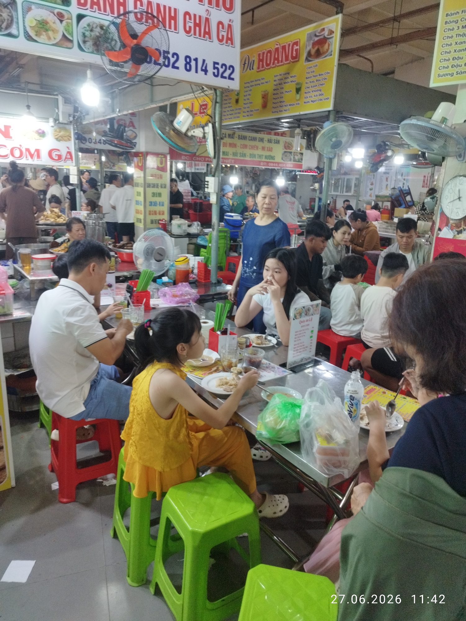 The image depicts a bustling outdoor food market in Phan Thiết, Vietnam, likely a popular local eatery given the signage and arrangement. In the foreground, a group of people are seated at metal tables, engaged in eating and drinking. A young girl in a yellow top and orange pants is prominently featured, eating from a plate. To her left, a man in a white polo shirt and blue jeans is also seated and appears to be eating. Other individuals, including adults and children, are visible around the tables, some interacting and others focused on their meals.
The environment is characterized by numerous food stalls and vendors, indicated by the colorful signs with Vietnamese text and images of food items. There are overhead fans to combat the heat, typical of such outdoor markets. Various food and drink items are visible on the tables, including plates of food, glasses, plastic bottles, and packaged items. The overall atmosphere suggests a lively and casual dining experience. The date stamp at the bottom right corner indicates "27.06.2026 11:42," suggesting the image was taken in the late morning or early afternoon.