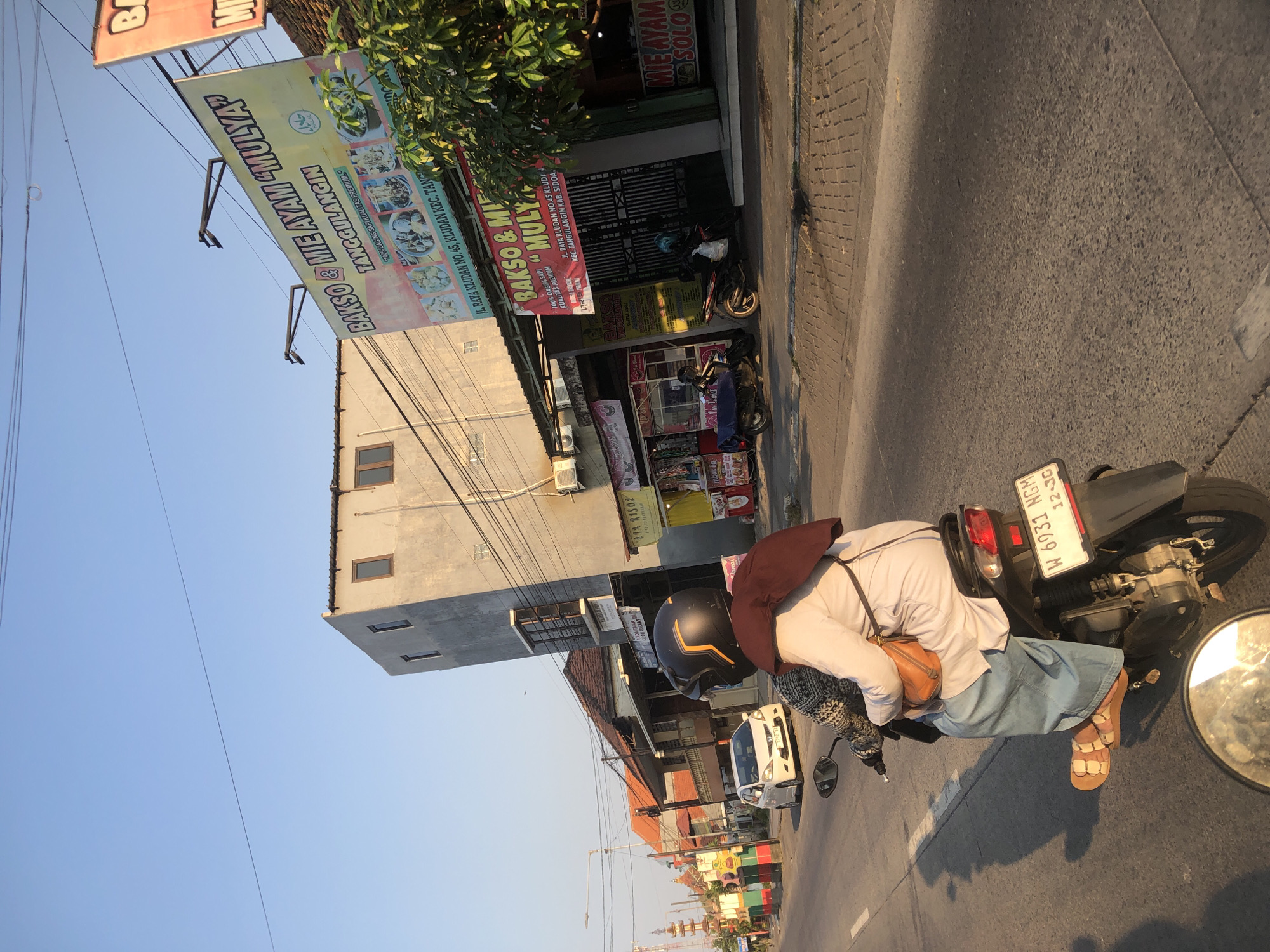 A still, eye-level shot captures a street scene in Boro, Indonesia, under a clear, pale blue sky during a period of late afternoon sun. The focus is on a motorcyclist in the foreground, seen from behind, wearing a helmet and a light-colored top with a brown headscarf and a brown purse. The motorcycle's license plate reads "W 6031 NGM". To the right of the motorcycle, a white car is visible on the road. The background features a row of buildings with a mix of architectural styles, including a tall, modern-looking grey structure with small windows and older, lower buildings with tiled roofs. Several street signs and advertisements are visible, including one for "BAKSO & MIE AYAM 'MULYA'". The road surface appears to be asphalt with a brick-patterned sidewalk on the right. The overall lighting suggests a warm, late afternoon or early morning light.