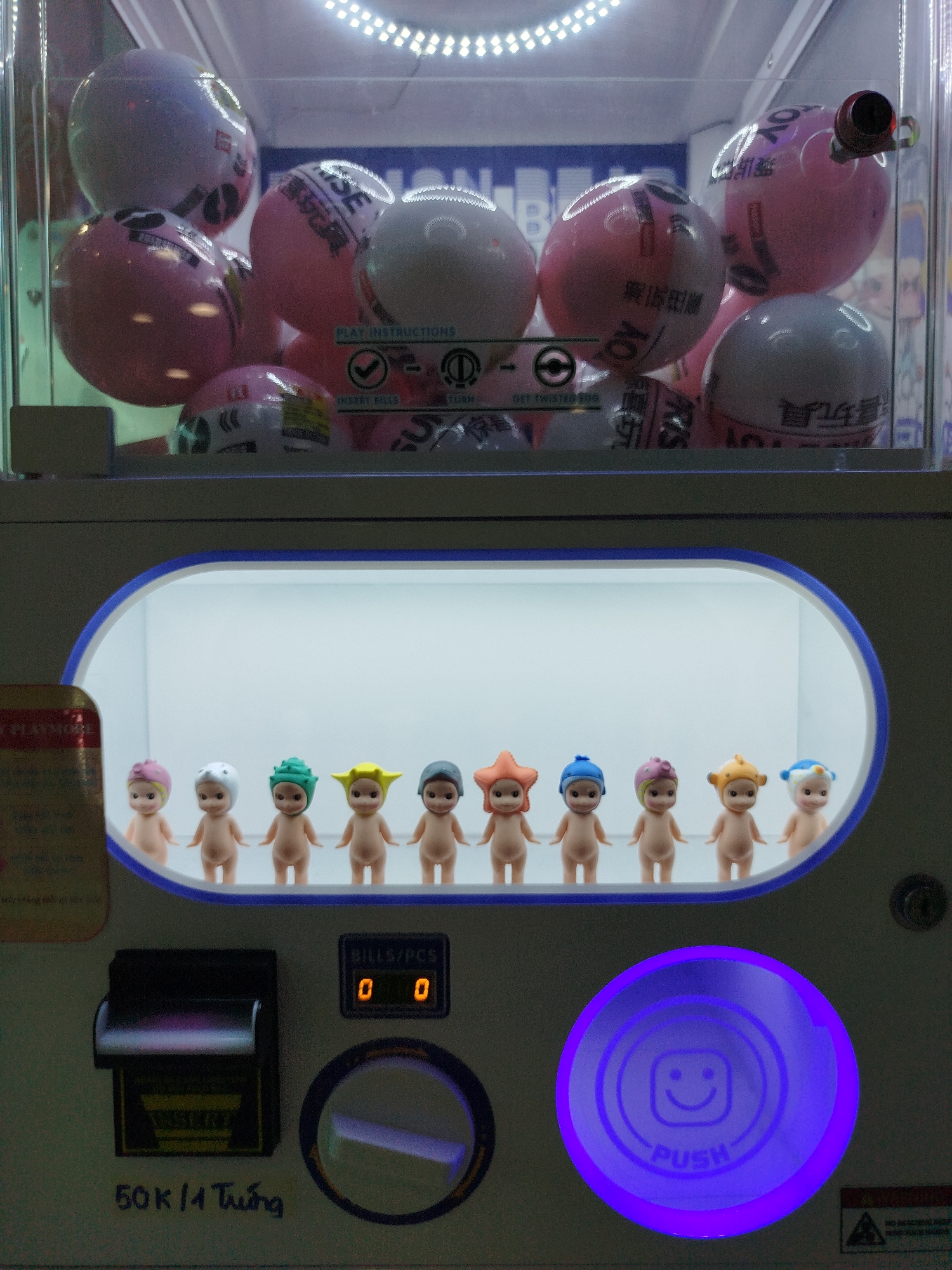 The image shows a toy vending machine, likely located in Da Lat, Vietnam, based on the price label of "50K/1 Trứng" (50,000 Vietnamese Dong per turn). The upper compartment of the machine displays various pink and white toy-filled capsules. Below, a lineup of small, naked doll figures with different colored headwear is visible behind a brightly lit display. The machine features a bill/coin insert, a twisting mechanism, and a large, illuminated "PUSH" button with a smiley face.