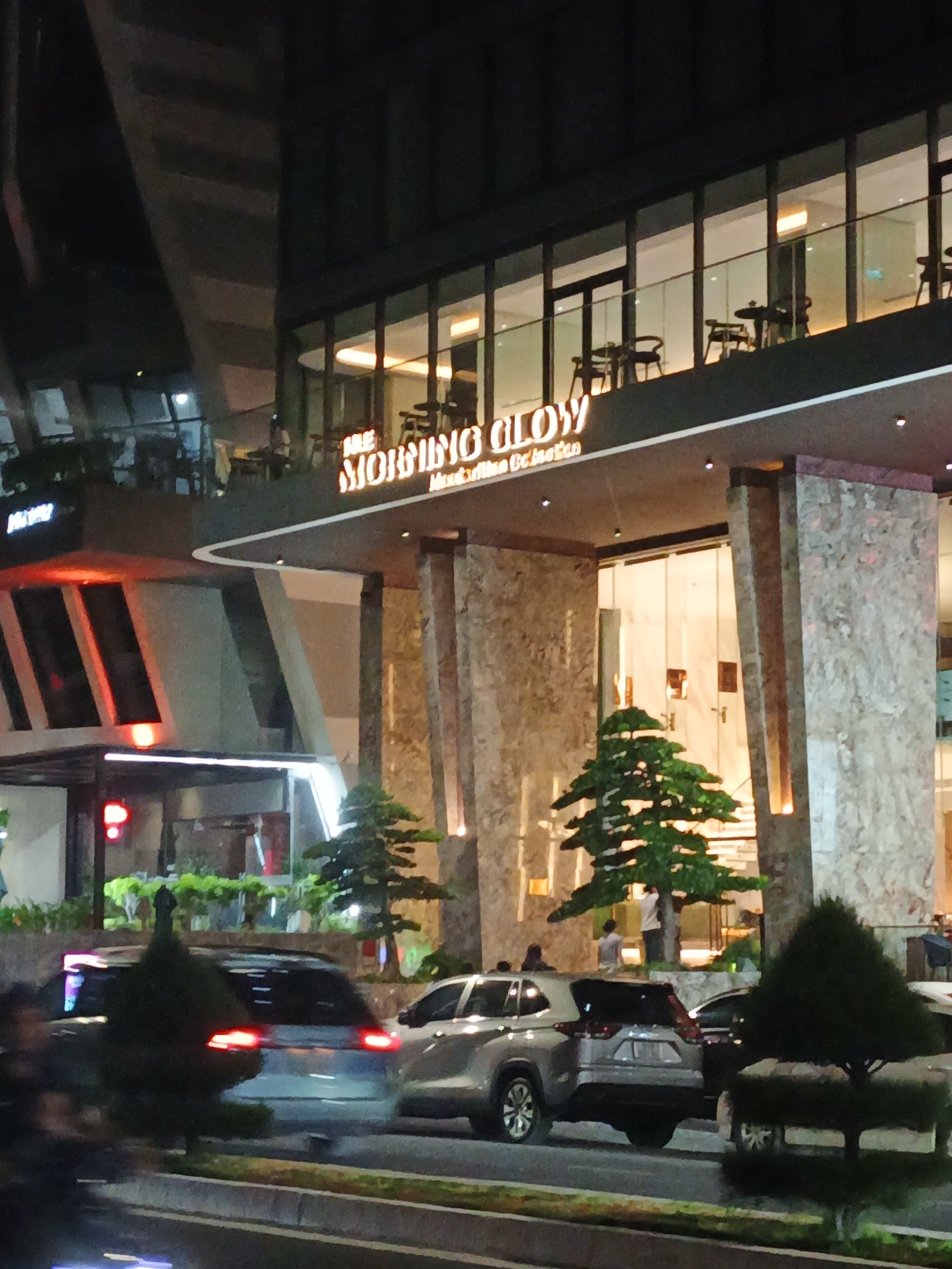 This is a nighttime, outdoor, medium-angle shot of a modern building's entrance, likely a cafe or restaurant, in Bac Nha Trang Ward, Vietnam. The building features a sleek, dark facade with large windows revealing an illuminated interior where tables and chairs are visible. A prominent neon sign above the entrance reads "THE MORNING GLOW" in stylized orange lettering, with smaller text below it that is illegible.
The entrance itself is framed by substantial pillars with a speckled, marble-like texture. In front of the building, two artistically shaped, dark green trees are placed, adding a touch of greenery to the urban scene. Beyond the trees, several cars are parked or driving on the street in front of the building. The streetlights and interior lighting create a warm glow, reflecting off the vehicles and the building's surfaces.
There are a few people visible: some are inside the building, and a couple are standing near the entrance. The overall atmosphere appears calm and inviting, suggesting a pleasant evening setting. The time of day is clearly night, with the artificial lights being the primary source of illumination. No specific weather conditions are evident, but the lighting suggests it is not raining.