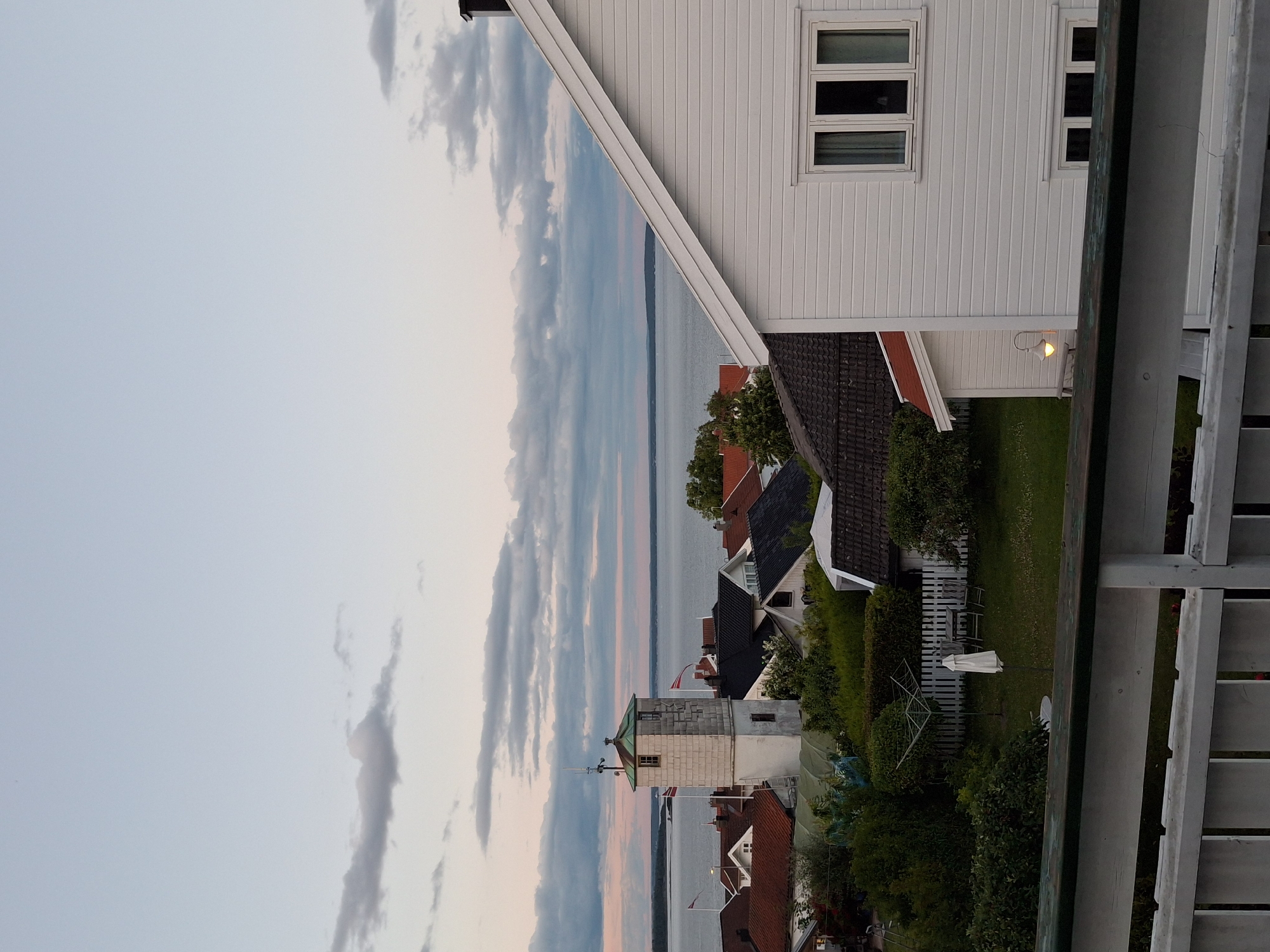 The image depicts a view overlooking Åsgårdstrand, Norway, at dusk. In the foreground, a white wooden railing with weathered green trim is visible. Beyond the railing, a residential area with several houses is spread out.
The houses feature a variety of roof colors, including red and dark gray. One prominent structure in the middle ground is a white, square tower with a dark green roof, resembling a lighthouse or a small observation tower. Two Norwegian flags are flying from poles near this tower.
The houses are surrounded by lush green trees and bushes. A neatly maintained lawn with a white picket fence is visible in the middle ground, with a clothesline and a closed white umbrella. In the foreground to the right, a section of a white, wooden house with horizontal siding and a visible window is present. A single outdoor lamp with a warm glow is also visible on this house.
In the background, a body of water, likely the Oslofjord, stretches towards the horizon. The sky above is a soft blend of pale blue and light pink, with scattered gray clouds, indicating the late evening light. The overall scene conveys a peaceful, serene coastal atmosphere.