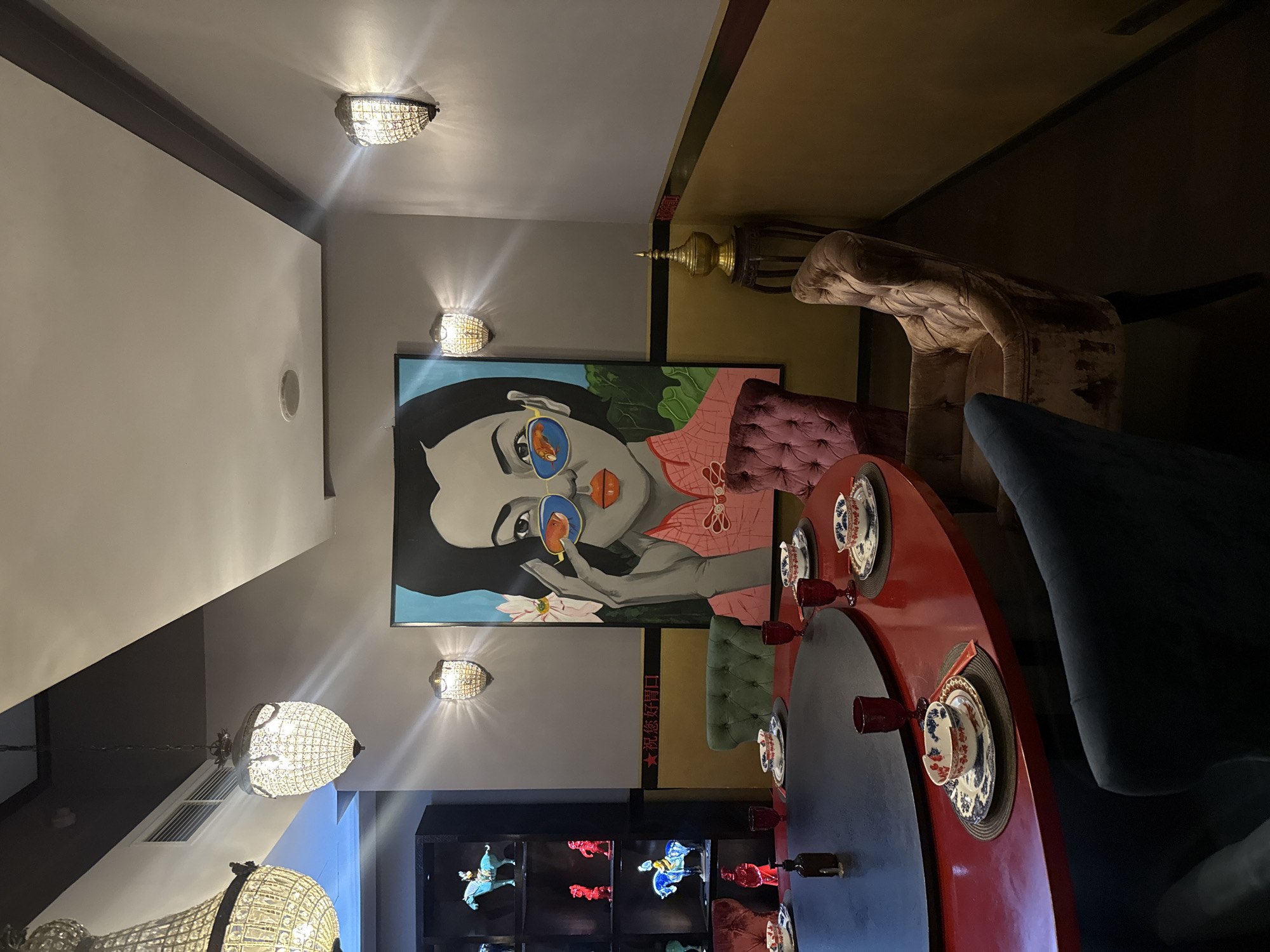The image depicts the interior of a dimly lit restaurant in Barvikha, Russia, furnished in a modern, eclectic style with East Asian influences. A large, circular red dining table dominates the foreground, set for a meal with blue and white patterned bowls, plates, and red-stemmed glasses. Dark blue and velvet chairs surround the table.
A striking, large-scale painting of a woman wearing sunglasses with fish reflected in the lenses hangs on the wall behind the table. The artwork is vibrant and features a color palette of blues, pinks, and greens. Two wall-mounted, ornate crystal sconces illuminate the painting and surrounding wall space.
To the left of the painting, a black shelving unit displays several decorative figurines of horses and people in red and blue, illuminated by spotlights. Above the dining area, two large, elaborate crystal chandeliers hang from the ceiling, casting a warm glow.
The walls are painted in a neutral gray, with a horizontal band of gold and red running along the lower section. In the background, a small, ornate golden urn or finial sits on a dark wooden stand. The overall ambiance suggests an upscale dining establishment with a focus on artistic decor.