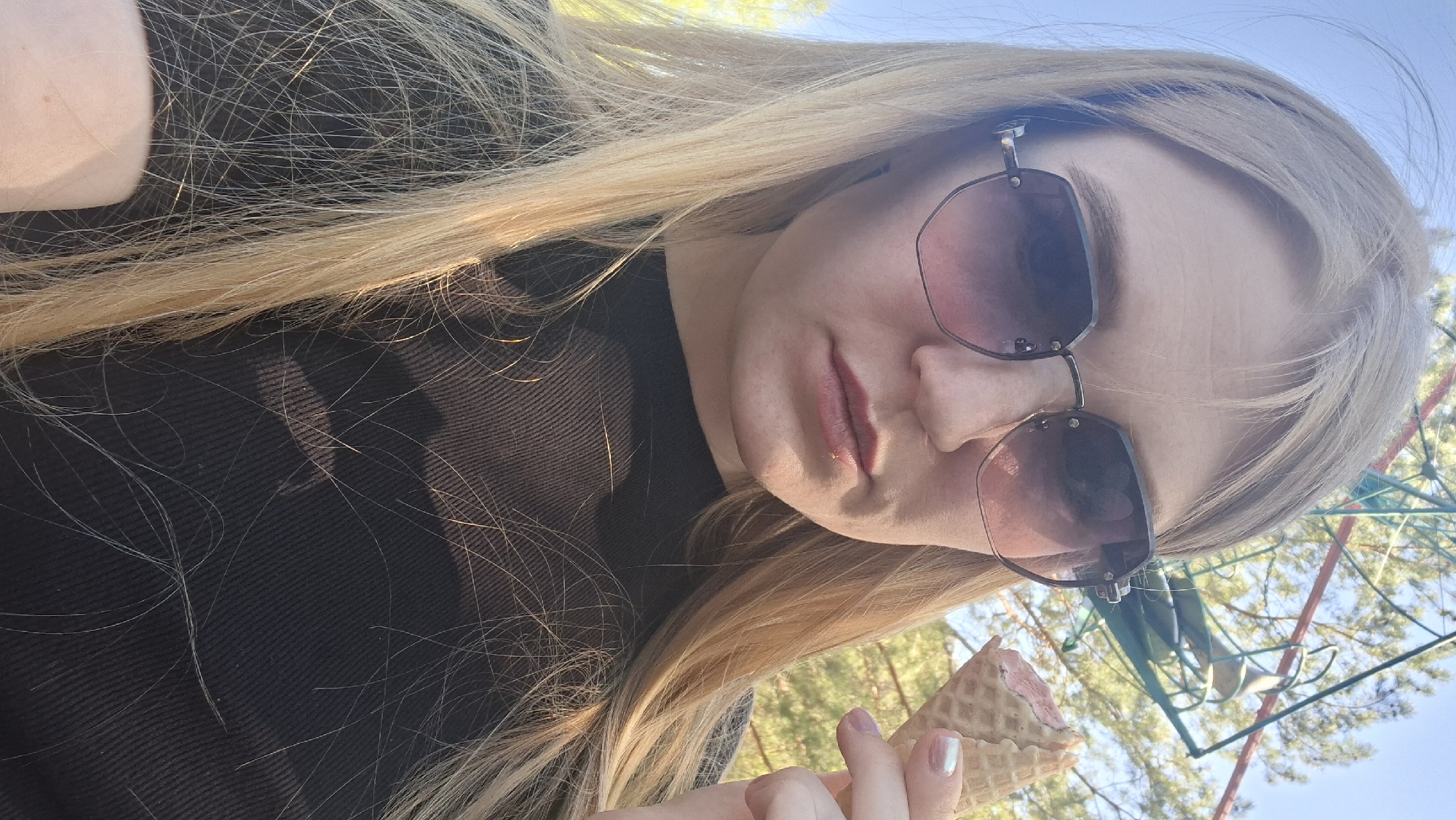 A woman with long blonde hair is holding an ice cream cone. She is wearing sunglasses and a dark top. The background shows trees and what appears to be a playground structure. The location may be in the city of Chaikovsky, Russia.