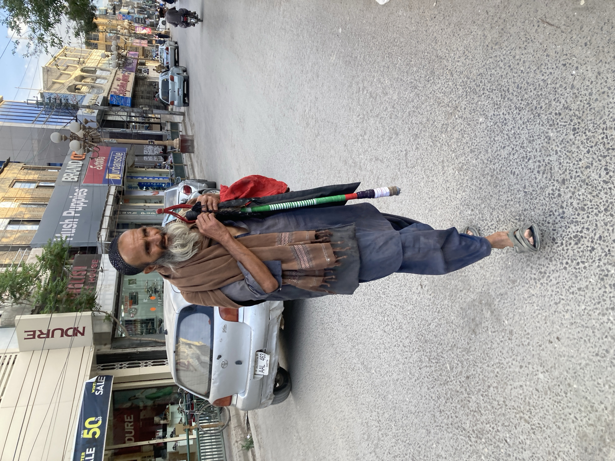 An older man, identified by the license plate "AAL 463" as being in Quetta, is standing on a city street. He is wearing a black skullcap, a brown shawl, and what appears to be a navy blue outfit, and gray sandals. He is carrying a long pole or walking stick over his shoulder, along with what appears to be a rolled-up umbrella and a red cloth. He is standing next to a silver compact car, which is parked on the street. The background shows shops and buildings, including signs for "Hush Puppies," "Brand," "Serwis," and "urbansole." There is also a shop with a "Winter Sale" banner. Other cars and motorcycles can be seen on the street.