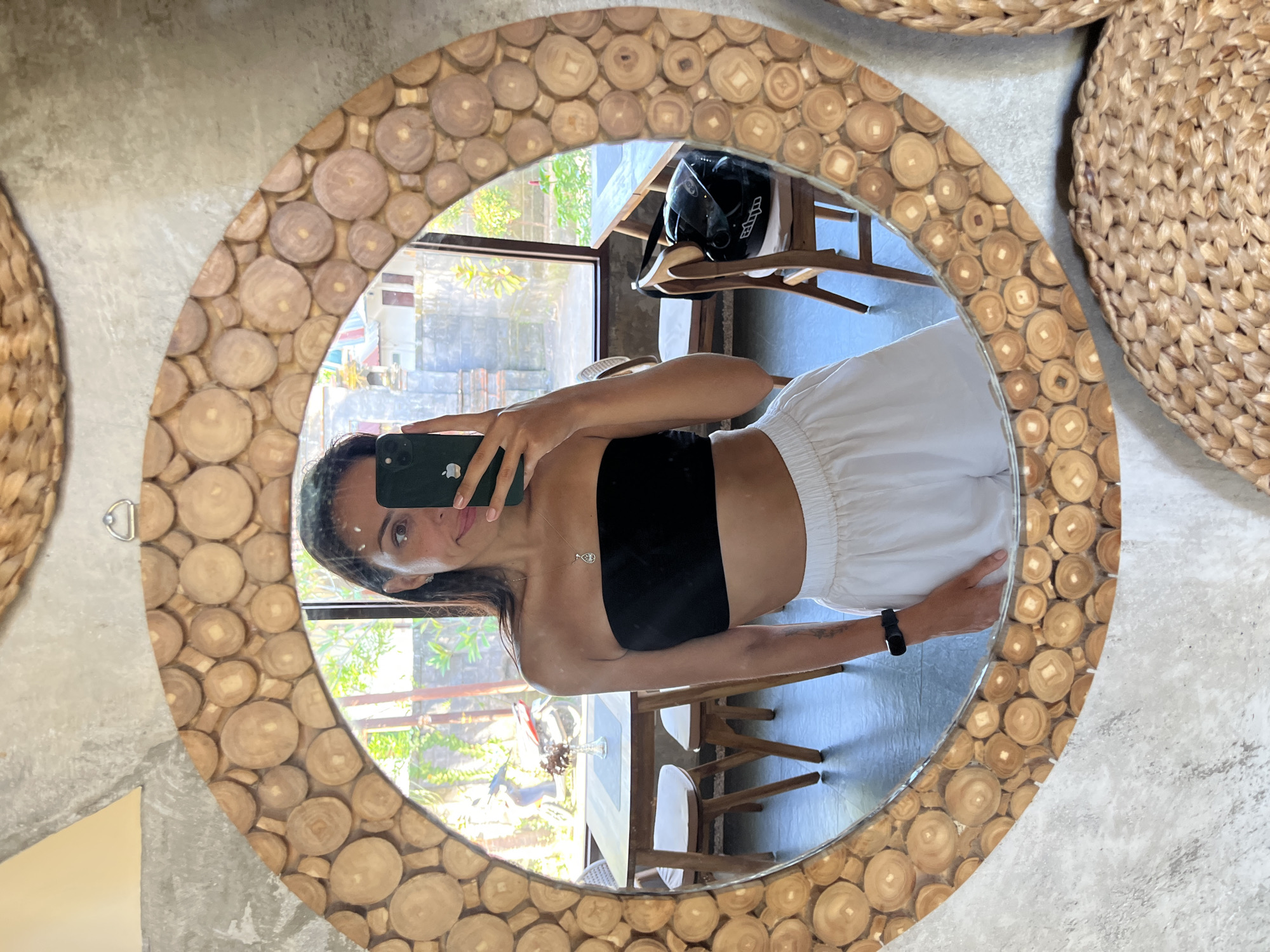 A woman takes a selfie in a round mirror made of small wooden disks. She wears a black tube top and white shorts. The reflection shows a room interior, including a table with chairs and a helmet on a shelf. Outside the window, greenery is visible, suggesting an outdoor or garden area. The mirror is hanging on a grey concrete wall next to woven baskets. Sukawati, Indonesia.