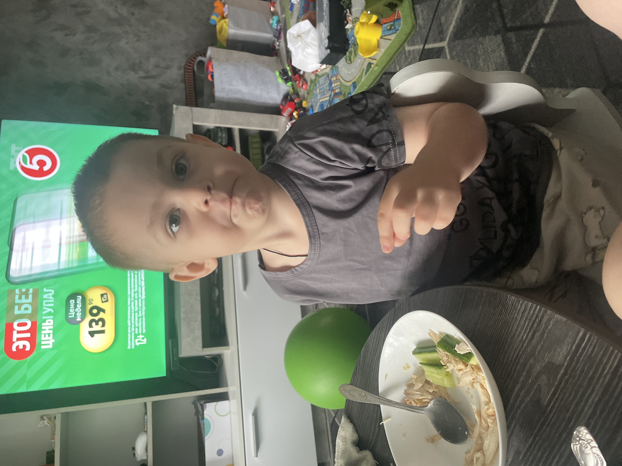 A young boy sits at a dark wooden table in a dimly lit room. In front of him is a white bowl containing shredded chicken and cucumber slices, with a spoon resting inside. To his left, a large, vibrant green ball sits on the table. The boy is wearing a grey t-shirt with a black graphic print on the sleeve and grey shorts. He is looking towards the camera with a slightly pursed expression. Behind the boy, a television screen displays a bright green advertisement for a sale, featuring text in Russian and a circular logo with the number 5. The background shows shelves with various items, including toy bins overflowing with colorful toys and what appears to be a gray textured wall. The lighting suggests this is an indoor residential setting, possibly a living room or dining area. There are no visible indicators to suggest the location is in Zhukovo, Russia.