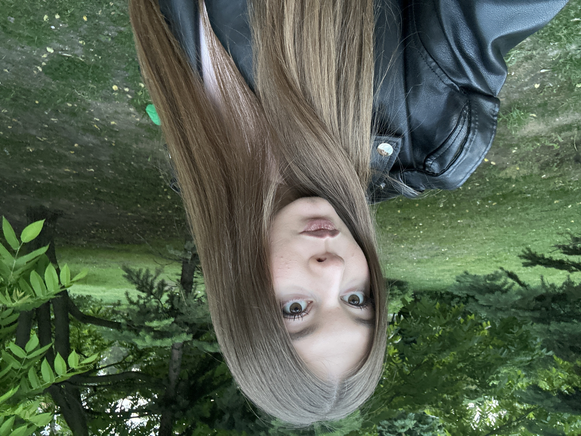 A close-up, chest-up portrait shows a young individual, likely female, against an outdoor, natural backdrop, suggesting a park or forested area in Kazan, Russia. The subject has long, straight, light brown hair that flows down, partially obscuring her shoulders and blending into the background. She is wearing a black leather-like jacket with stitching details and a visible silver snap button on the lapel, over a faint white inner garment.
Her face is positioned towards the upper center of the frame, with her forehead, prominent brown eyes, nose, and slightly parted lips clearly visible. She gazes directly into the camera with a wide-eyed, perhaps surprised or intently focused expression.
The background is rich with various shades of green, consisting of lush grass, trees with bright green deciduous leaves, and darker green coniferous trees. Some bare ground or a path can also be seen. The lighting suggests it is daytime, likely a soft, ambient light without harsh shadows. There is no visible text in the image.