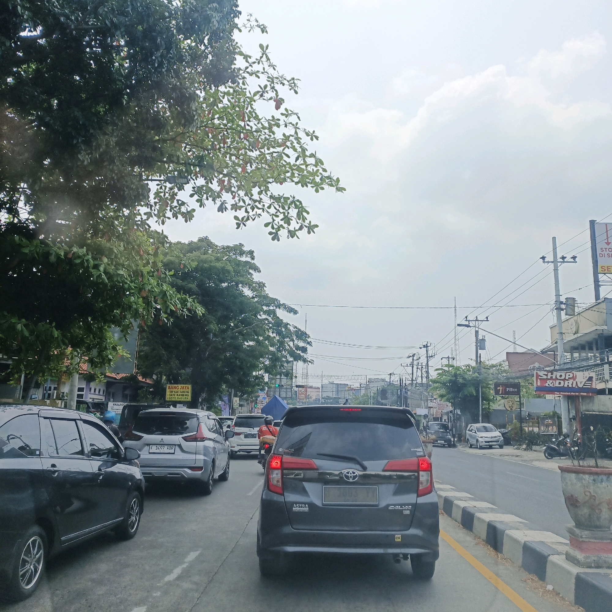 The image captures a street scene in Semarang, Indonesia, characterized by moderate traffic and urban infrastructure. In the foreground, a dark-colored car is visible from the rear left, parked on a paved road. To its right, a silver MPV (Multi-Purpose Vehicle) with license plate F 1497 ABR is seen from the rear, also on the road. Further ahead, a dark gray MPV with license plate H 1003 YY is in traffic, its brake lights illuminated. The road is lined with trees on the left, with dense green foliage and some reddish-brown leaves. Buildings and shops are visible along the right side of the road, including signage for "BANG JAYA," "SHOP & DRIVE," and "PIBee." Several utility poles with electrical wires crisscross the sky, typical of an urban environment. The sky is overcast with scattered clouds. In the distance, a tall communication tower can be seen. A motorcycle with a rider wearing an orange jacket is also present in the traffic flow. The road has yellow and black painted dividers along the curb. The overall impression is a typical daytime scene on a busy street in Semarang.