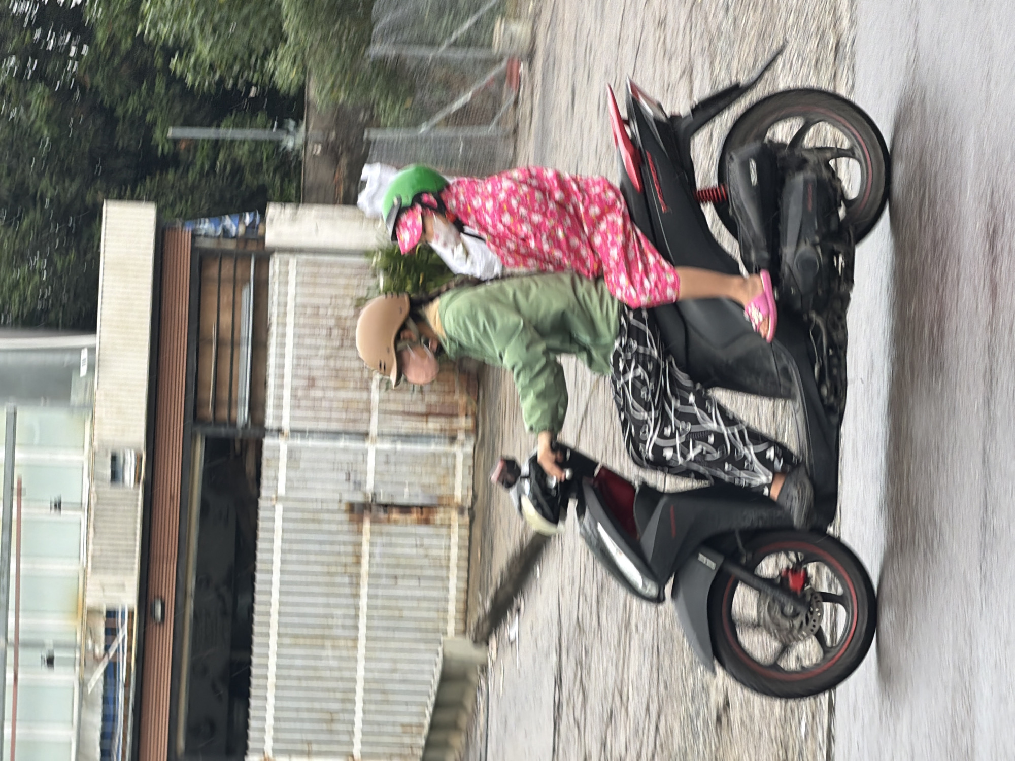 Two individuals, likely female, are riding on a dark-colored scooter on a wet, paved street. The person in the front is wearing a light green jacket, black and white patterned leggings, and a light brown helmet with a pink face mask. The person in the back is wearing a pink and white floral print full-body covering, a green helmet, and pink sandals. Both individuals appear to be wearing face masks. The scooter has red accents on the wheels and a red rear seat rest. The background shows a corrugated metal building with a large opening, possibly a garage or storefront, and some foliage. The scene is captured from a side profile, with a slight motion blur indicating the scooter is in motion. The environment suggests an urban setting in Nha Trang, Vietnam, possibly during or after rainfall given the wet road surface.