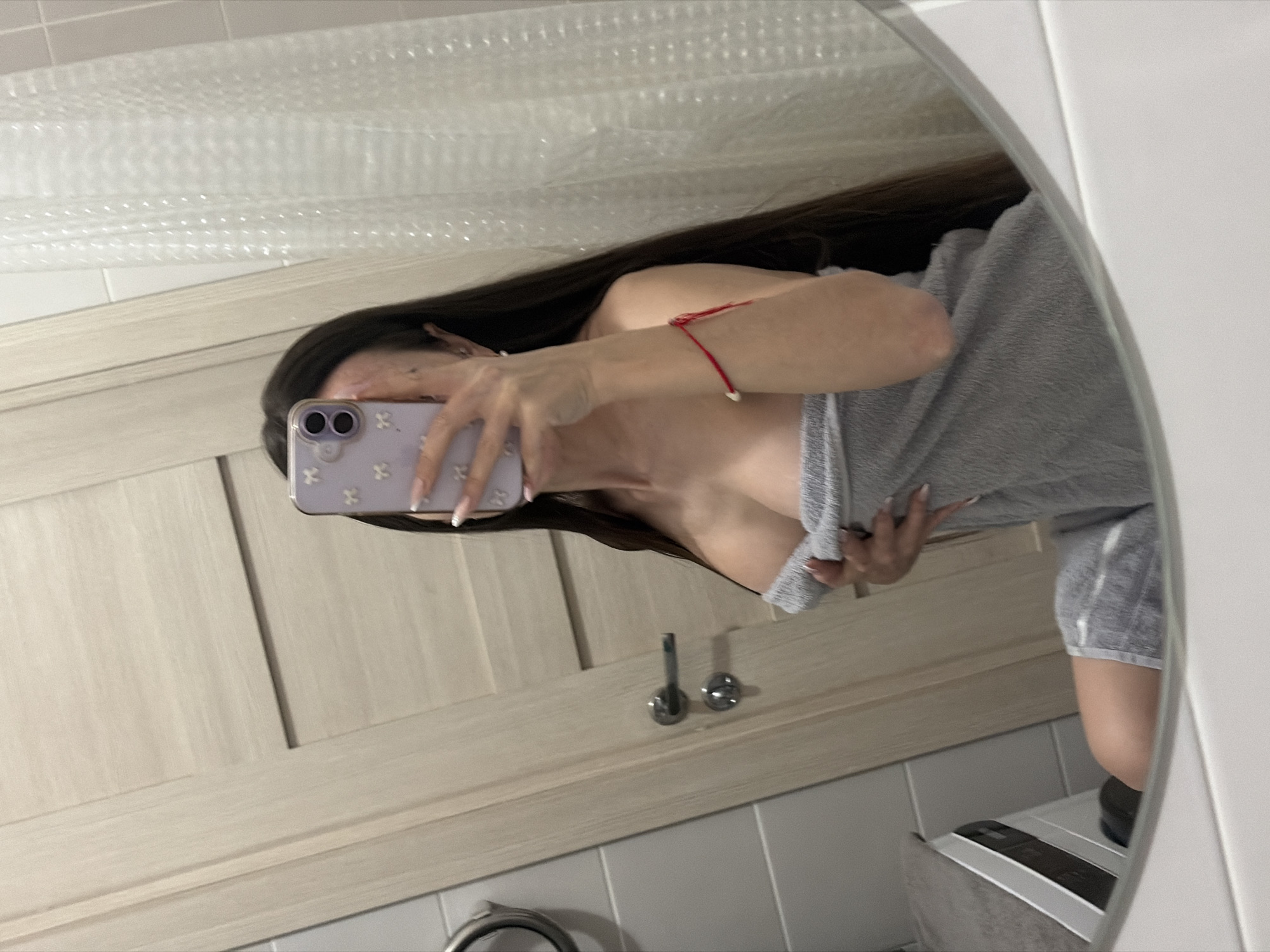 A mirror selfie captures a person in a bathroom, with the image rotated 90 degrees clockwise. The individual, likely female, has long dark hair and is wrapped in a grey, textured towel, suggesting they have recently showered or bathed. Their face is obscured by a light purple smartphone with a case featuring small white patterns, which they hold with their right hand, revealing long, light-colored manicured nails. A thin red string bracelet is visible on their left wrist.
The reflection in the oval mirror reveals a bathroom interior. Above the person's head, a crinkled, clear material, possibly a shower curtain liner with a bubble-wrap texture, is visible. Below the person in the reflection are light wood-grained cabinet doors or paneling, with two silver faucet handles or knobs reflected below the smartphone. White tiled walls are seen at the top and bottom edges of the mirror's reflection. In the bottom right corner, part of a white and grey appliance, resembling a washing machine or dryer, is reflected. The lighting appears to be artificial, typical of an indoor setting. No specific time of day or emotions are discernible. No visible text is present in the image. The scene takes place in a private bathroom in Saint Petersburg, Russia.