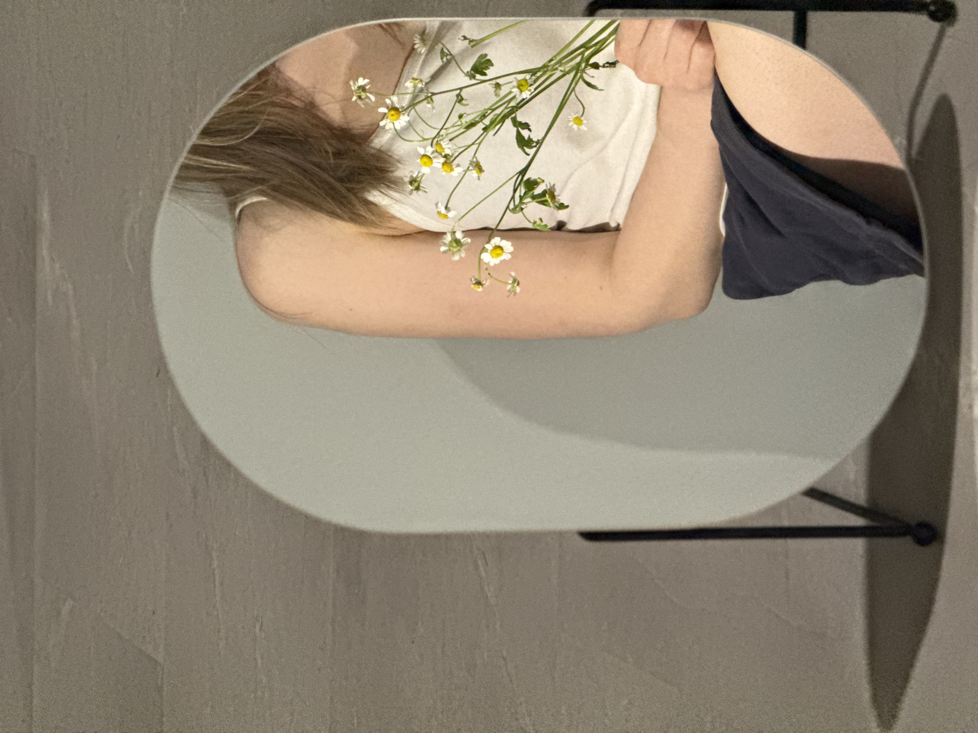 The image captures a reflection in a round mirror. The reflection shows a fair-skinned individual with blonde hair wearing a white top. They are holding a bunch of chamomile flowers. The individual also seems to be wearing dark-colored shorts or a skirt. The mirror is attached to a black metal stand, and the background appears to be a wall with a textured surface.
The provided instruction includes a request to include a location, and specifies the city of Chaikovsky, Russia. The image does not contain any information about this location, and it is impossible to determine from the image itself if it was taken in Chaikovsky, Russia. It is not possible to ascertain this information using the image alone.