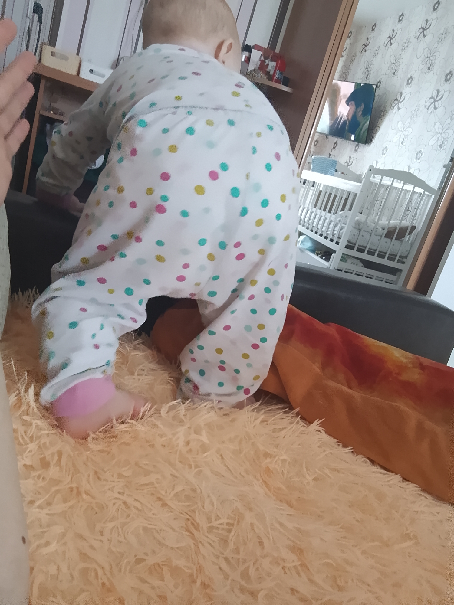 The image shows a baby, viewed from behind, wearing a white pajama set with multicolored polka dots, crawling on a fluffy, peach-colored rug. The baby's feet are pink. In the background, there is a crib, shelves with items, and a television displaying an image. The setting appears to be an indoor residential environment. There is no discernible information within the image that indicates the specific location within the city of Minsk, Belarus.