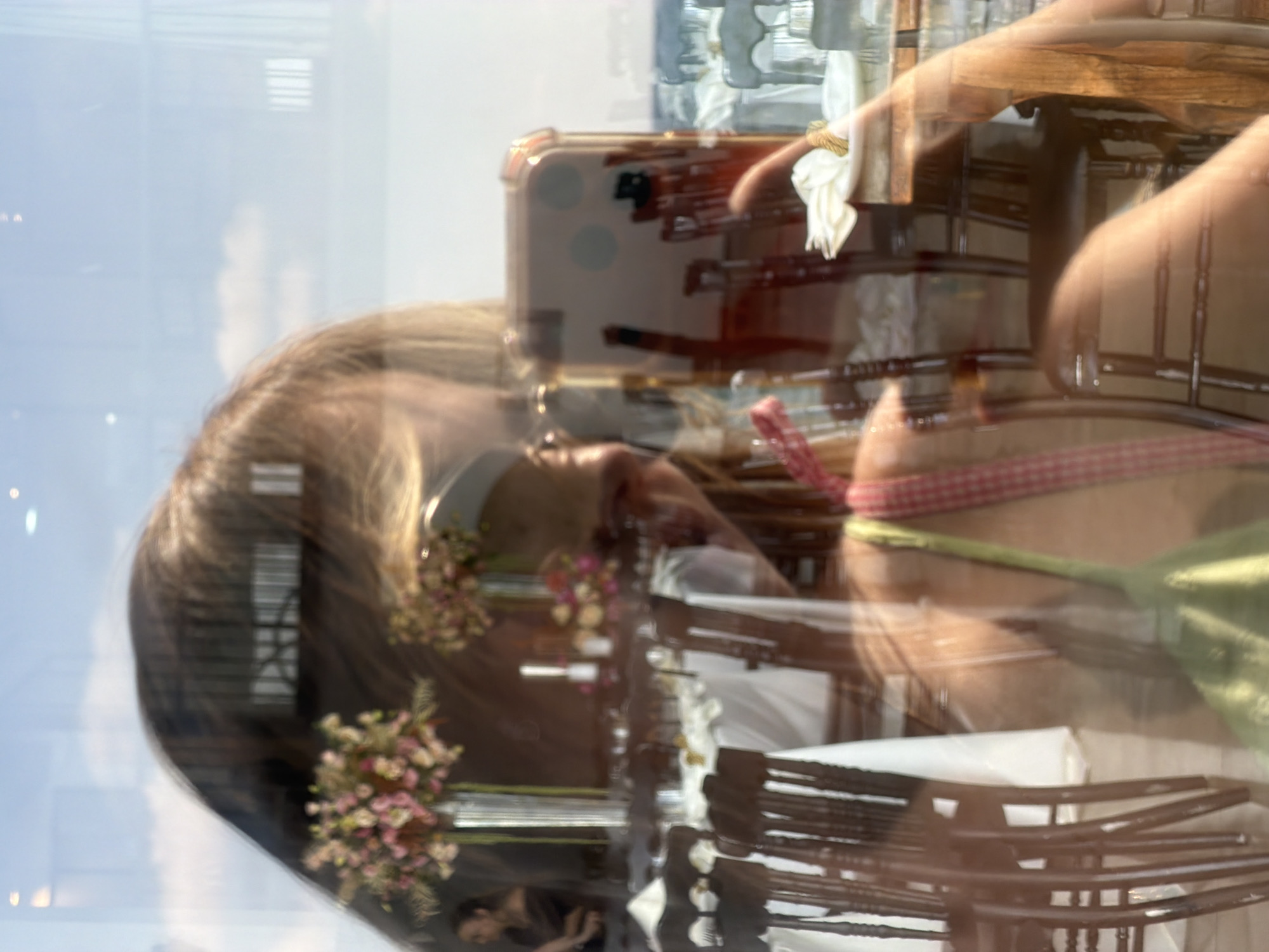 This is a reflection of a person on a glass surface. The person is facing the glass, and their phone is visible in their reflection, held in their hand as if taking a selfie. The reflection also shows a dining area with tables and chairs set up, suggesting a restaurant or event venue. In the background, through the glass, there are elements that appear to be the sky and possibly distant buildings. The presence of flowers on the tables suggests a celebratory or formal occasion. The lighting indicates it is daytime. The scene is set within Tel Aviv, Israel, though specific landmarks are not identifiable from the reflection alone. The reflection is slightly distorted and blurred, typical of images captured through glass.