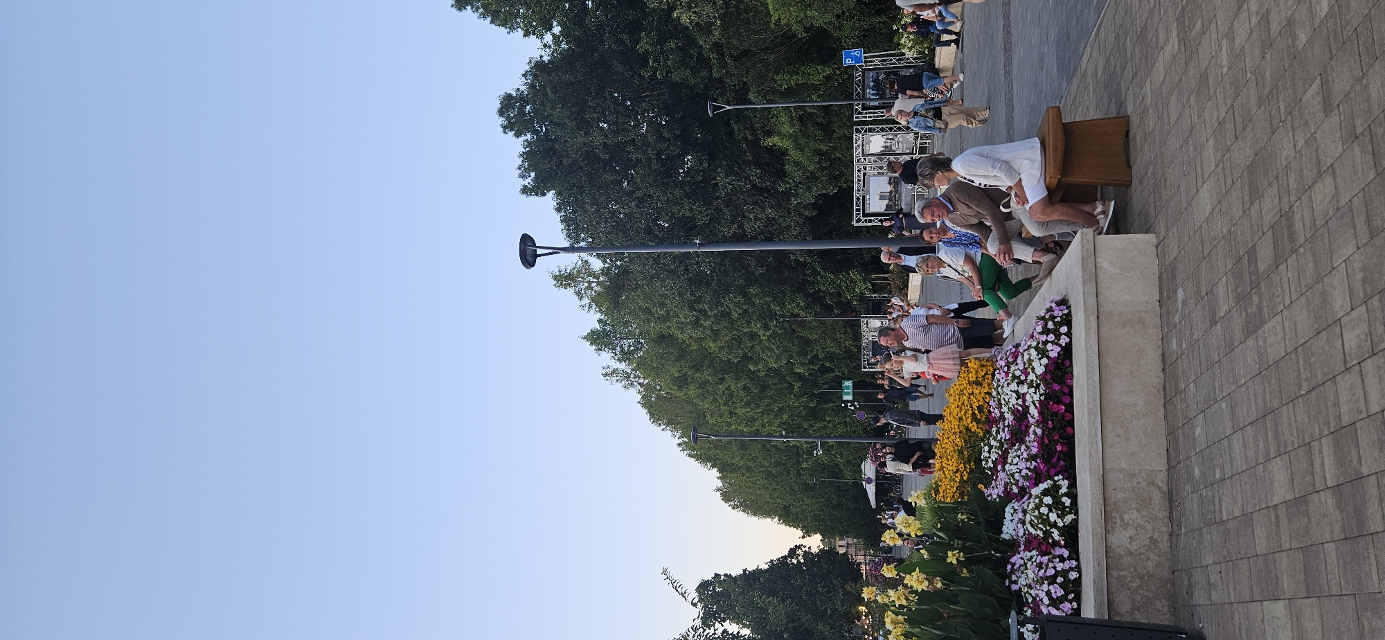 The image shows an outdoor scene in Hévíz, Hungary, likely a public square or park. In the foreground, a raised flower bed is filled with yellow, pink, and white flowers. Several people are seated on a wooden bench in front of the flower bed, facing towards the right of the frame. The people appear to be casually dressed and engaged in conversation. Behind the bench and flower bed, a paved walkway extends into the background, lined with trees and streetlights. Other individuals are visible walking and standing along the walkway and in the distance, suggesting a public gathering or event. Several large framed displays, possibly exhibits or information panels, are set up along the walkway in the middle ground. The sky is clear and blue.