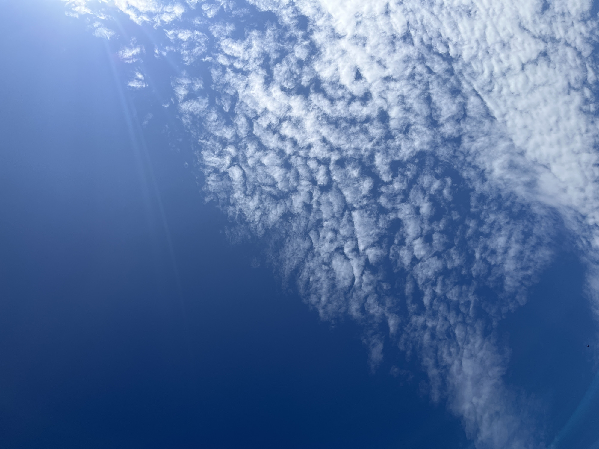 The image shows a bright blue sky with scattered white clouds. The clouds are in various shapes and sizes, with some appearing wispy and others more defined and lumpy. There is a bright light source, likely the sun, visible in the upper right corner, creating a lens flare across the sky. The sky appears to be clear of any structures or significant atmospheric phenomena. The location is identified as บ้านกาญจน์กนก 23, Thailand, suggesting an outdoor scene in that region.