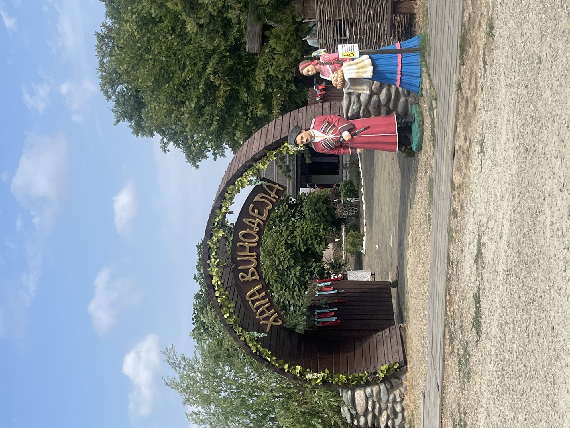 The image depicts an outdoor scene in Taman', Russia, featuring a rustic wooden archway decorated with greenery and grapevines. Gold lettering on the arch reads "ХАТА ВИНОДЕЛА," which translates to "Winemaker's Hut." Flanking the entrance to the archway are two life-sized mannequins. On the left, a male mannequin wears a traditional red Cossack-style coat with ornate trim and a black hat, holding a dagger. On the right, a female mannequin is dressed in a pink blouse and a vibrant blue skirt with pink trim, holding a basket of bread. The ground is a mix of gravel and wooden planks, and in the background, lush green trees and a building with a thatched roof are visible. The sky is blue with scattered white clouds.