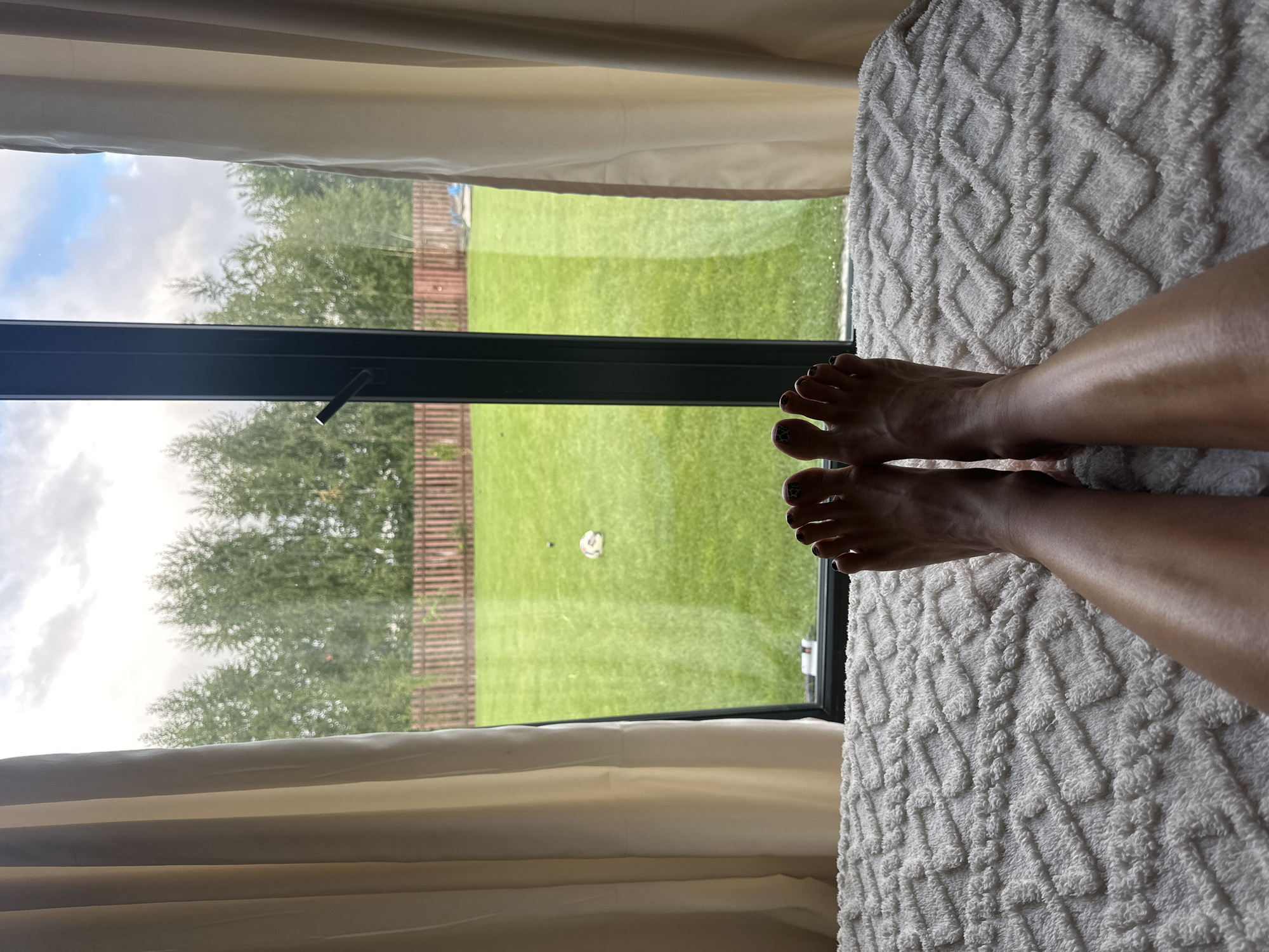 A first-person perspective shot shows a person's legs and feet resting on a white textured surface, likely a bed or couch, with a large window in the background. Through the window, a green lawn with a wooden fence and trees is visible. The sky above is cloudy. The person's feet have painted toenails.