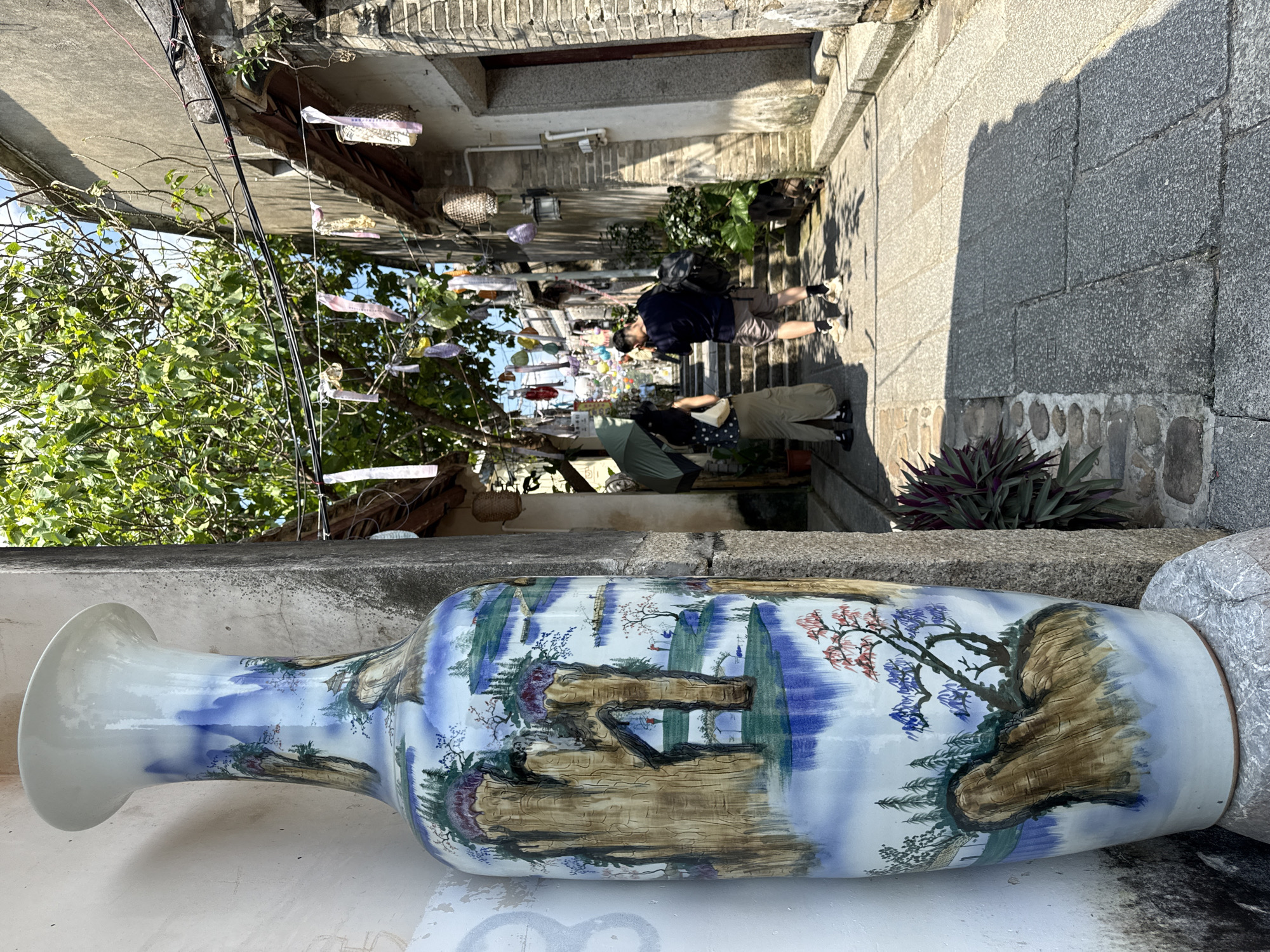 A tall, ornate porcelain vase painted with a landscape scene stands in the foreground on the left, leaning against a textured wall. The vase depicts mountains, trees, water, and figures in a traditional Chinese art style. To the right, a narrow, stone-paved alleyway recedes into the distance, lined with traditional brick buildings. The alley is decorated with colorful streamers and small woven baskets hanging from overhead wires. Two people are visible in the alleyway: a woman holding an umbrella, and a man with a backpack walking away from the camera. The scene is illuminated by bright sunlight, casting shadows on the ground. The overall impression is one of a historic or culturally significant area within the city of Longgang District, China.