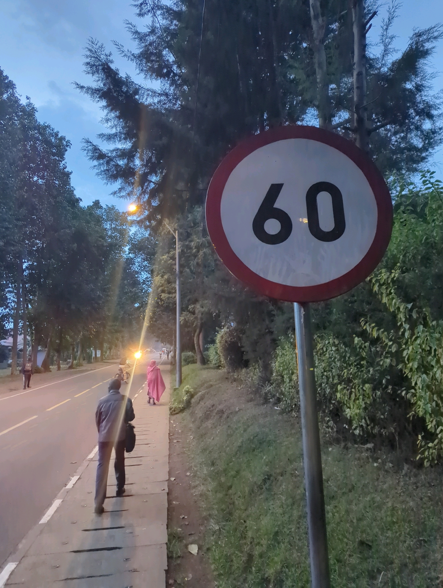 This image shows a roadside scene in Musanze District, Rwanda. A speed limit sign indicating "60" is prominently displayed on the right side of the frame. In the foreground, a man in dark clothing walks away from the camera on a paved sidewalk. A woman in a pink garment is visible further down the sidewalk, walking towards the right. A cyclist is also present on the road. The road itself has lane markings and appears to be a two-lane street. Trees line both sides of the road, and there is vegetation along the sidewalk. Streetlights are visible, illuminating the scene. The sky is overcast, suggesting it might be dusk or early evening. In the background, there are more trees and some indistinct buildings or structures.