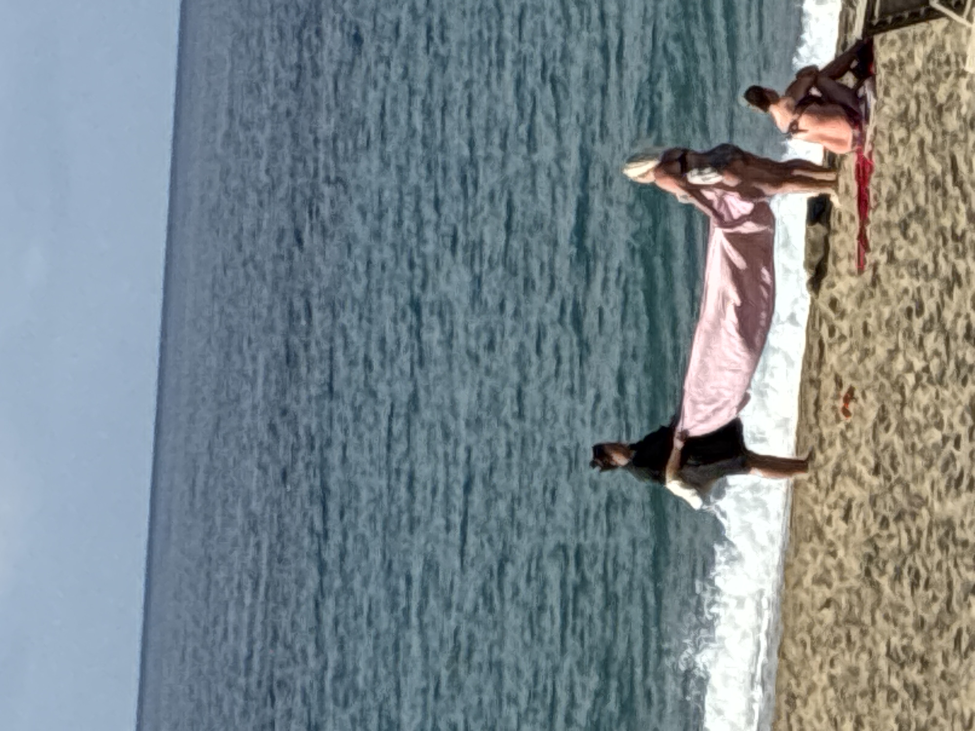 This image depicts a beach scene with the sea in the background and a sandy shore in the foreground. Three individuals are present. In the middle of the frame, two women are standing, with one holding a large pink towel. The woman on the left is wearing a dark top and shorts, while the woman on the right is wearing a bikini. To the right of the standing women, a third person, likely a man, is seated on the sand, also in swimwear. Waves are visible crashing on the shore. The environment is consistent with a beach in Alanya, Turkey, characterized by its coastline and sandy beaches.