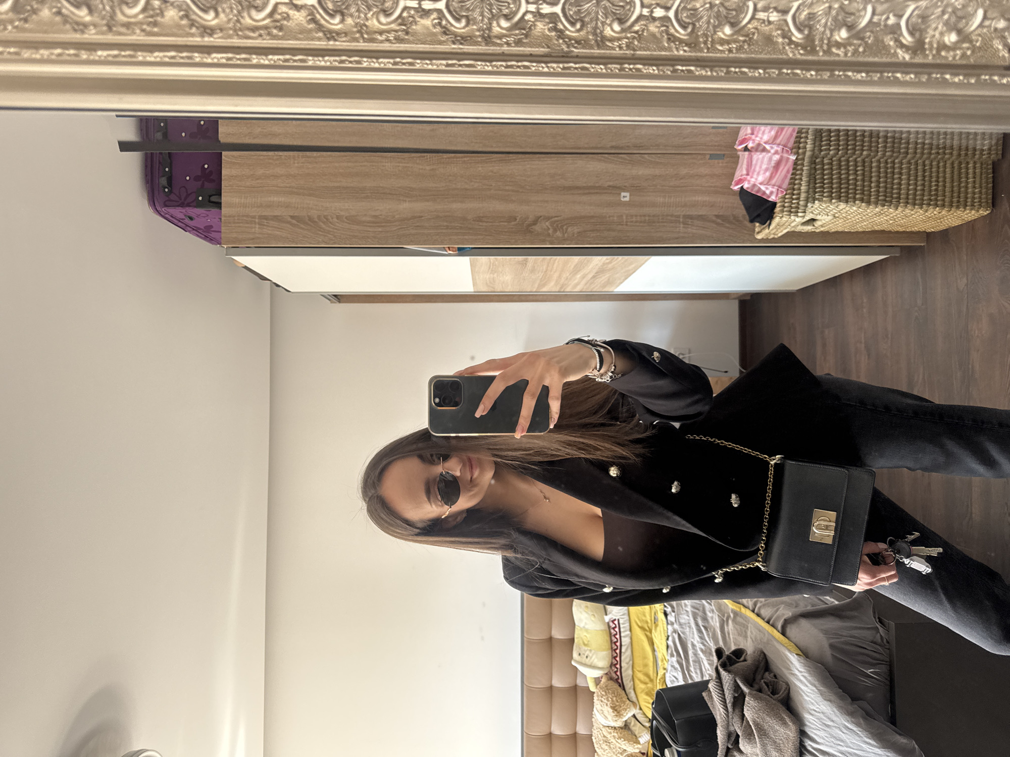 A woman is taking a selfie in a mirror. She is wearing a black blazer, black jeans, and sunglasses. She is holding a black purse. The background includes a bed with a gray comforter, a brown headboard, and a wardrobe. It's impossible to determine the exact location in Sofia, Bulgaria, without additional information.
